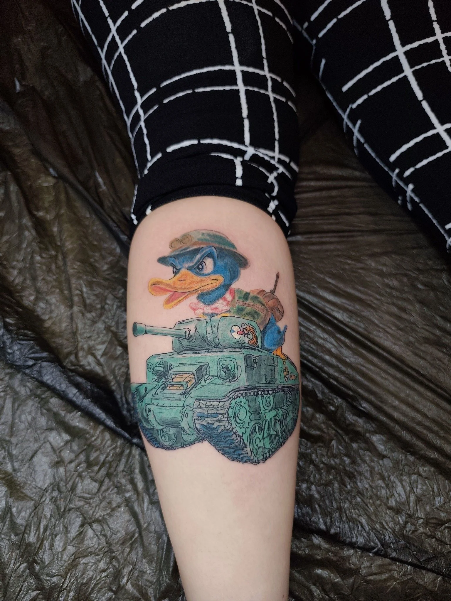 A colorful tattoo of a Duck dressed as a soldier or military figure, sitting on a military tank, on a person's lower leg.