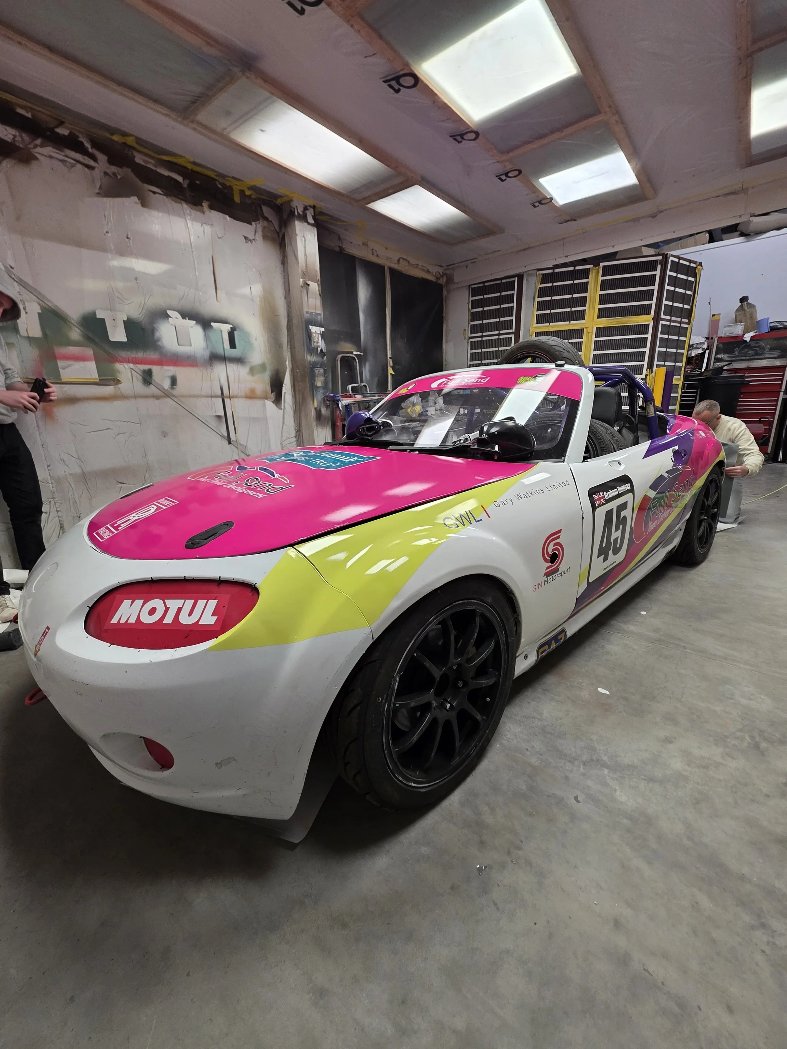 A race car with a white, pink, and yellow paint scheme in a garage or workshop, with tools and equipment around.