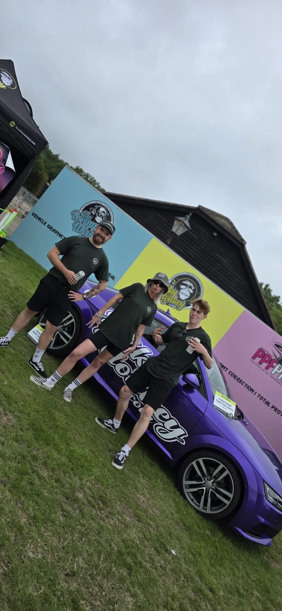 Three people stand beside a purple car with a large colorful advertisement in the background. They are wearing black T-shirts and shorts, and are outdoors on grass.