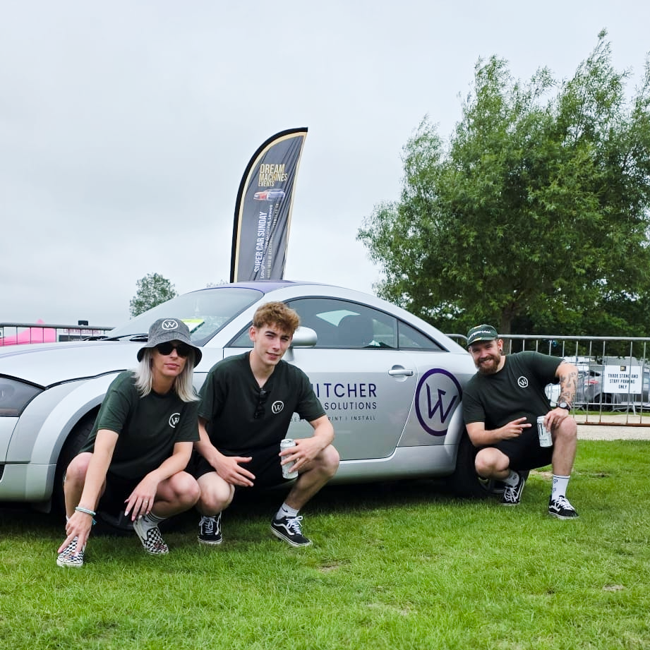 Three people crouching next to a silver car with VTCHER SOLUTIONS branding, outdoors on grass, with a tree and a fence in the background and a flag that reads 'DREAM MACHINES EVENTS'.