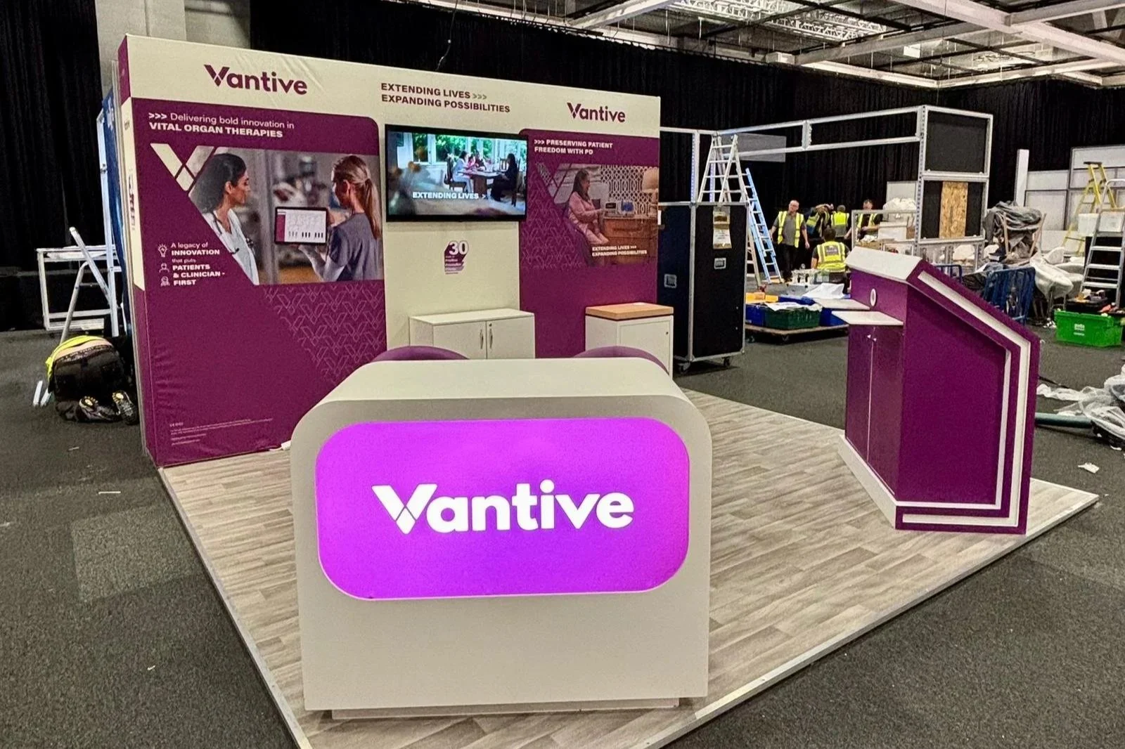 Exhibition booth for Vantive with purple and white color scheme, set up in an indoor event space. The booth has a reception desk with a glowing purple sign displaying 'Vantive', back panels with promotional material, and a small television screen. There are construction workers and equipment visible in the background.