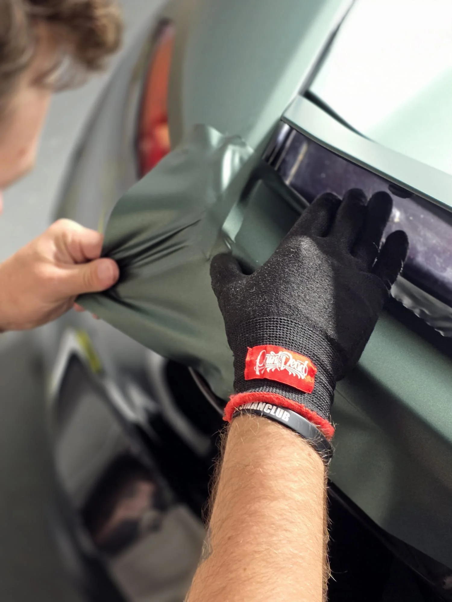 A person wearing black gloves and a wristband is applying a vinyl wrap to a car's surface. The image is taken from a close angle showing the hand pressing the wrap onto the car.