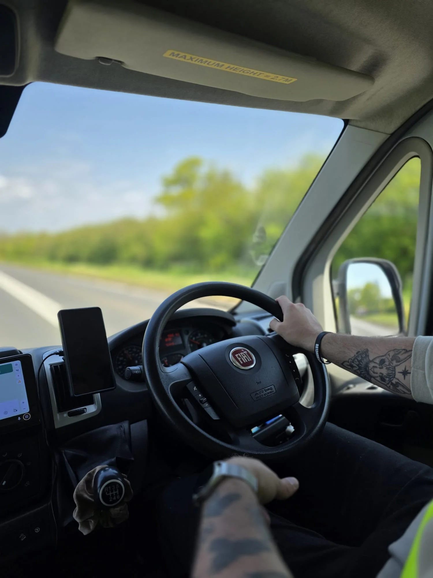 Inside a vehicle, the dashboard, steering wheel with Fiat logo, and driver's arm with tattoos are visible. Outside, a sunny day with blue sky and green trees seen through the windshield.