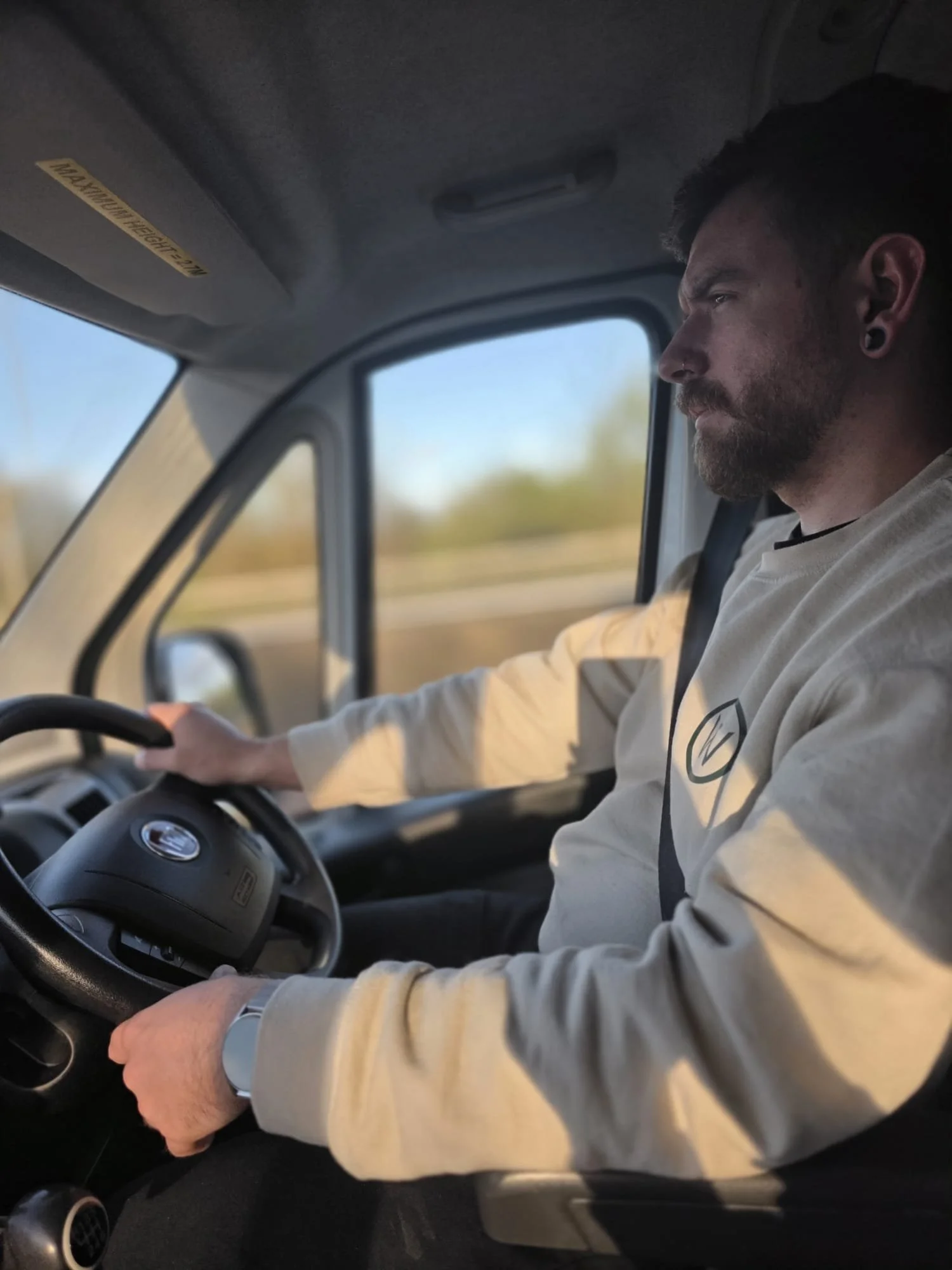 A man with a beard, dark earrings, and a light-colored shirt driving a vehicle, with a scenic view of trees and sky visible through the window.