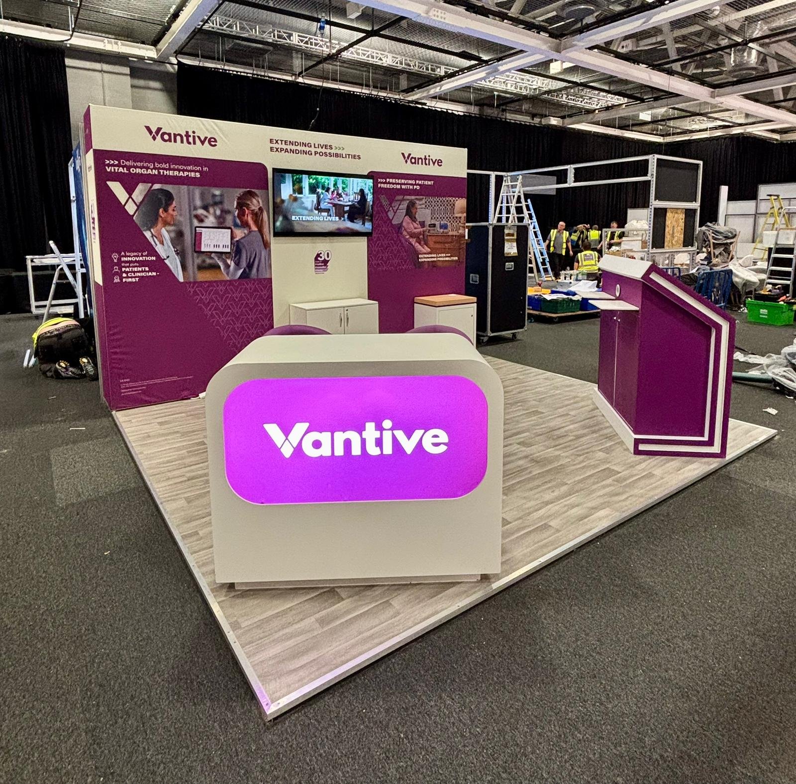 An exhibition booth for Vantive with a purple and white color scheme, featuring signs and screens discussing vital organ therapies, extending lives, and preserving patient freedom, with a small setup area and staff members in the background under a large indoor ceiling.