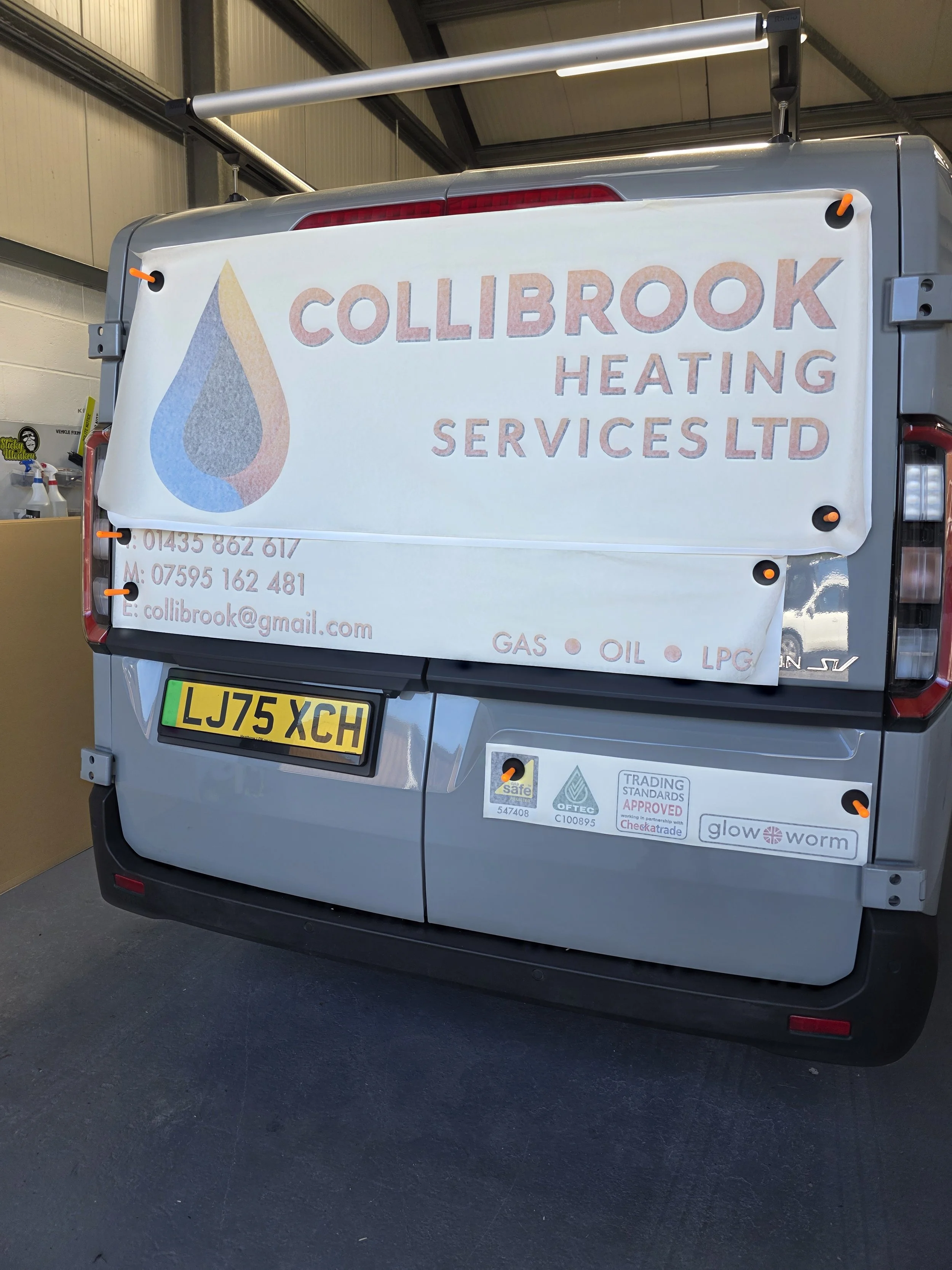 The back of a silver vehicle with a large sign for Collibrook Heating Services Ltd attached to it. The sign features a logo with a water droplet and contact information. Several stickers are on the vehicle, and a license plate reading 'LJ75 XCH' is v