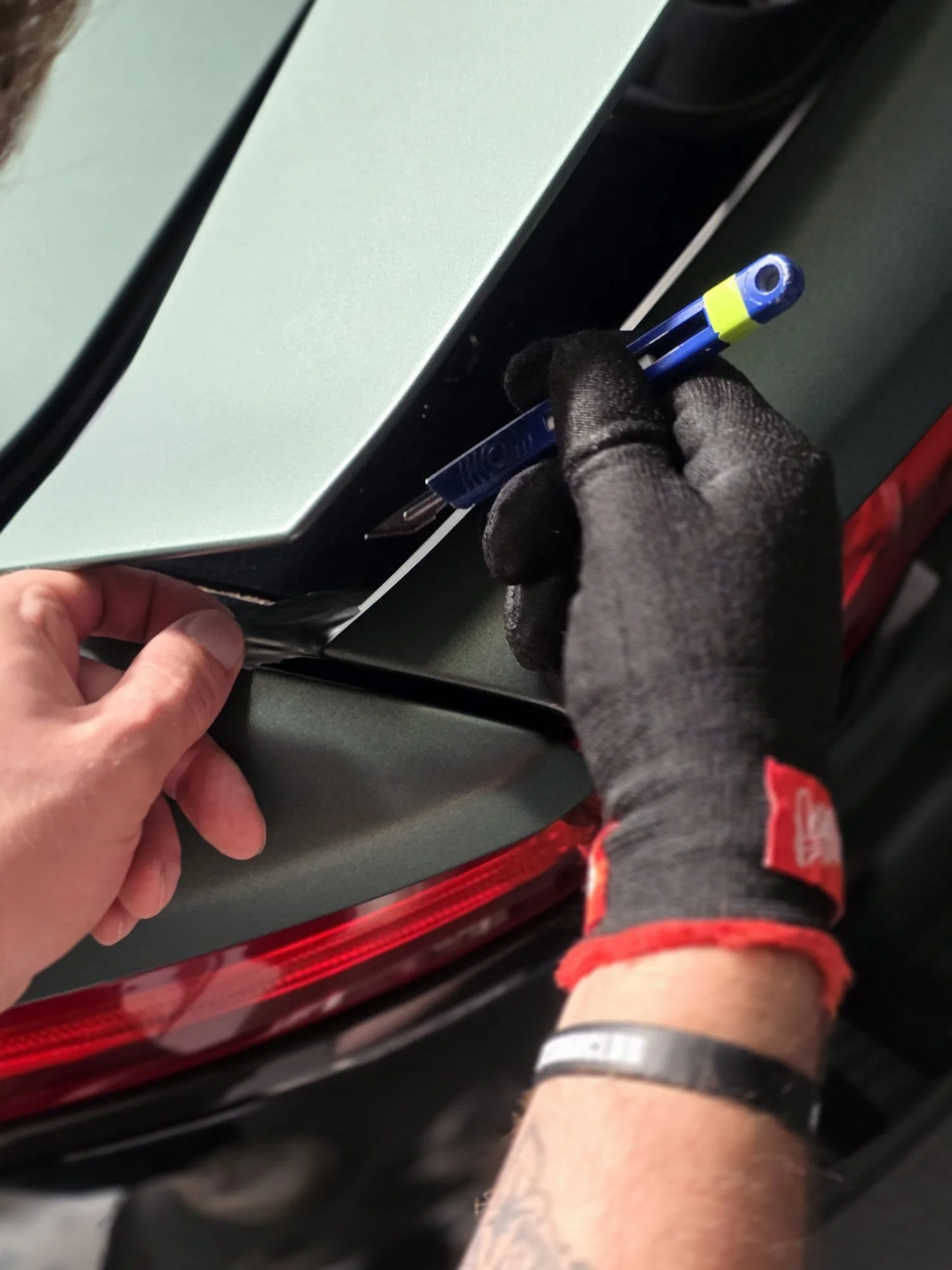 A person wearing black gloves is installing a blue and yellow automotive fuse into a fuse box in a vehicle. The vehicle's trunk lid and red taillight are visible.