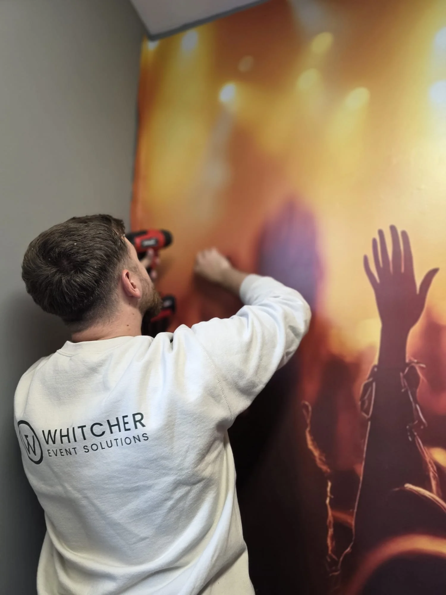 A man with brown hair and a beard using a power drill to attach something to a colorful abstract wall art with silhouettes of people reaching upward and lightning bolts.