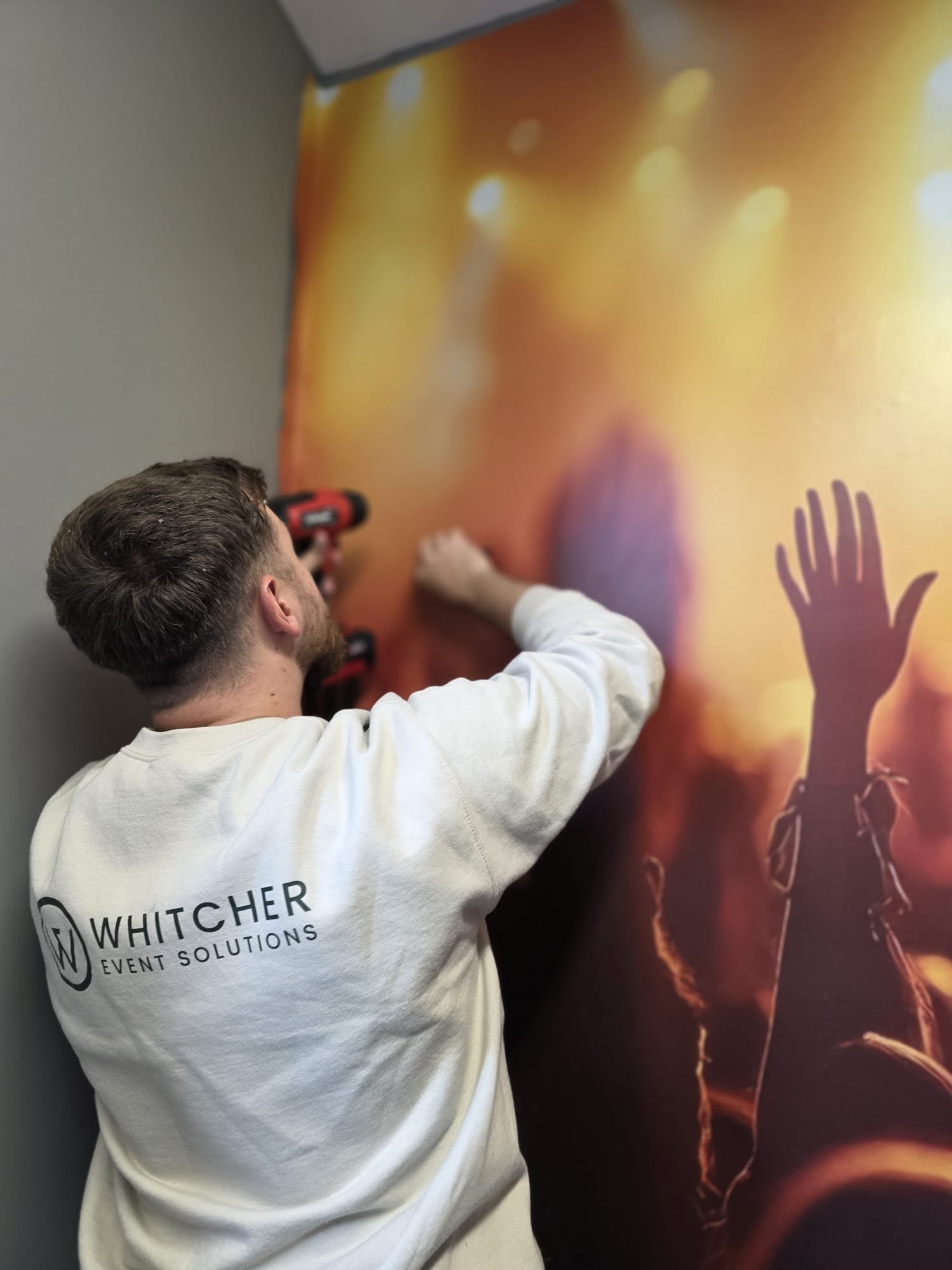 A man with a white shirt that has the logo 'WHITCHER SOLUTIONS EVENT SOLUTIONS' on the back, using a cordless drill to attach a poster or banner to a wall. The poster has an abstract image with orange and yellow hues and some dark outlines.