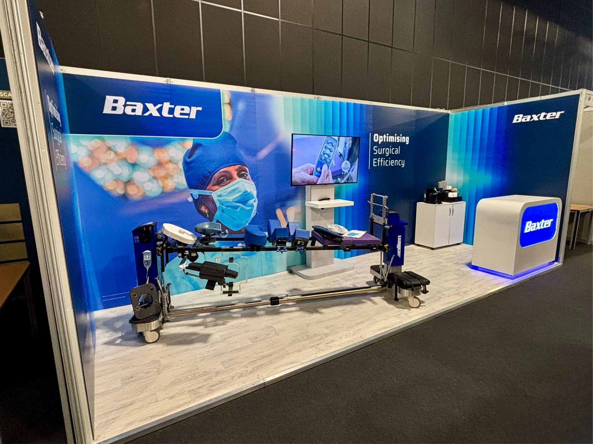 Trade show booth for Baxter, featuring medical equipment including a hospital bed and surgical tools, with a large banner showing a healthcare professional wearing a mask and surgical cap. The booth emphasizes optimizing surgical efficiency.