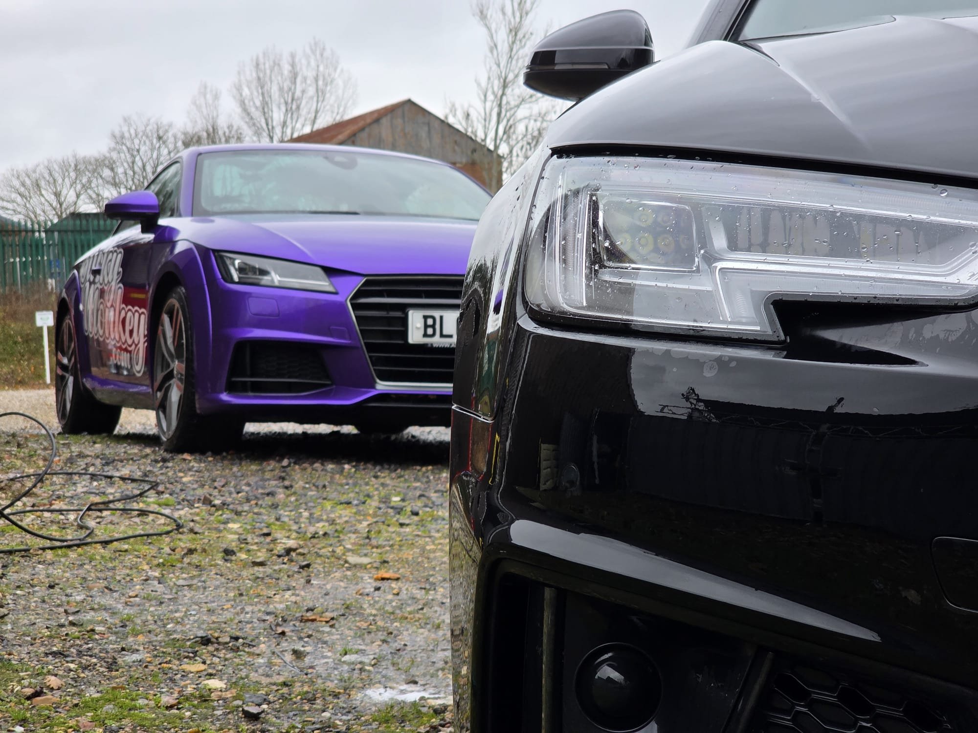 Close-up of the front of a black car with an updated headlight, with a purple car in the background parked on a gravel surface.