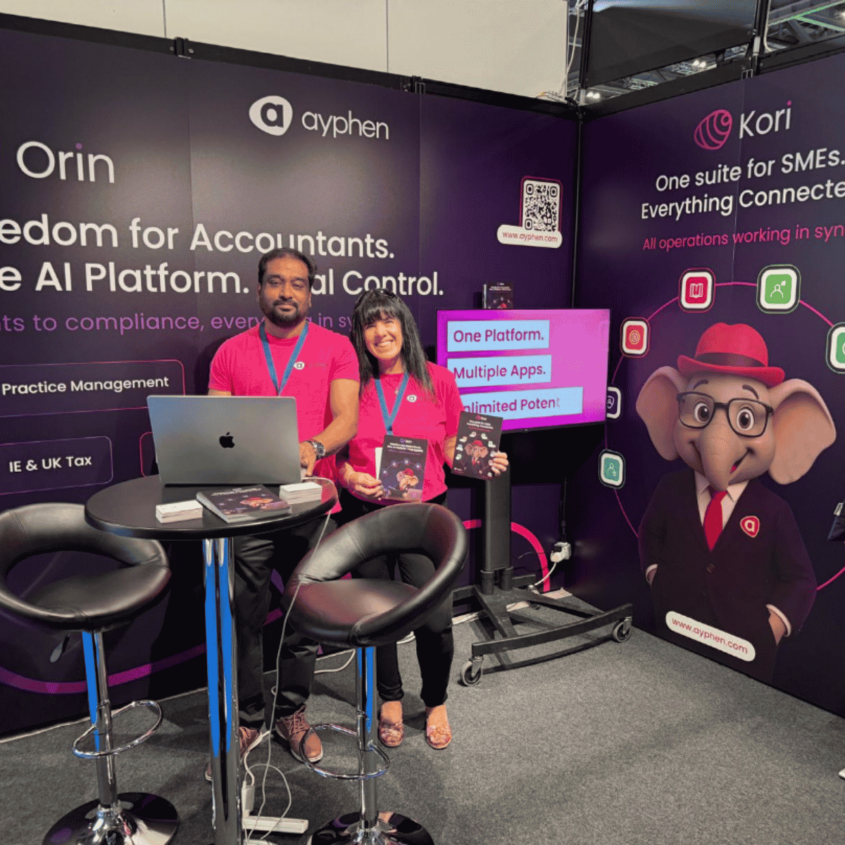 Two representatives at a booth for a company named Ayphen, promoting their AI platform for accountants. The booth has large banners with information, including a cartoon elephant mascot wearing glasses and a red hat. The representatives are wearing matching pink shirts, standing behind a table with a laptop and flyers, smiling at the camera.