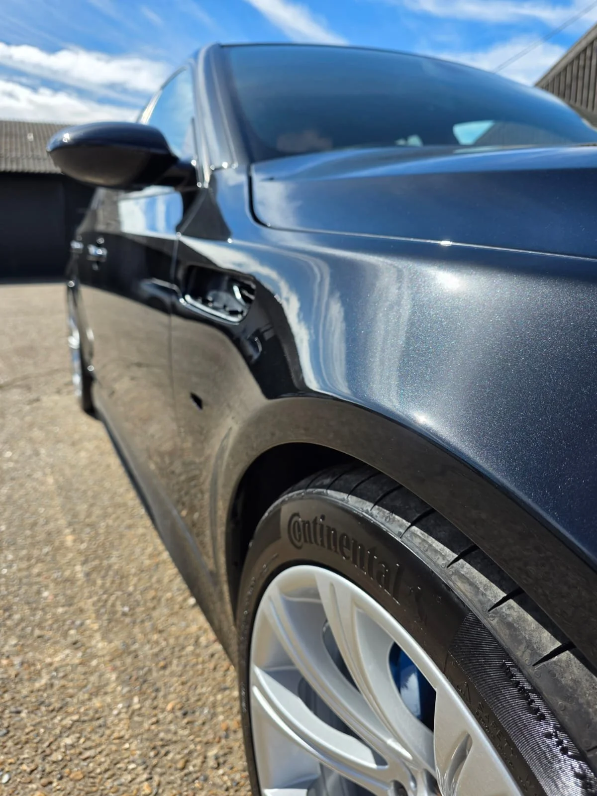 Close-up of a dark blue car's front side, showing the wheel with a Continental tire, glossy paint, side mirror, and a partly visible door handle, with a warehouse and blue sky in the background.