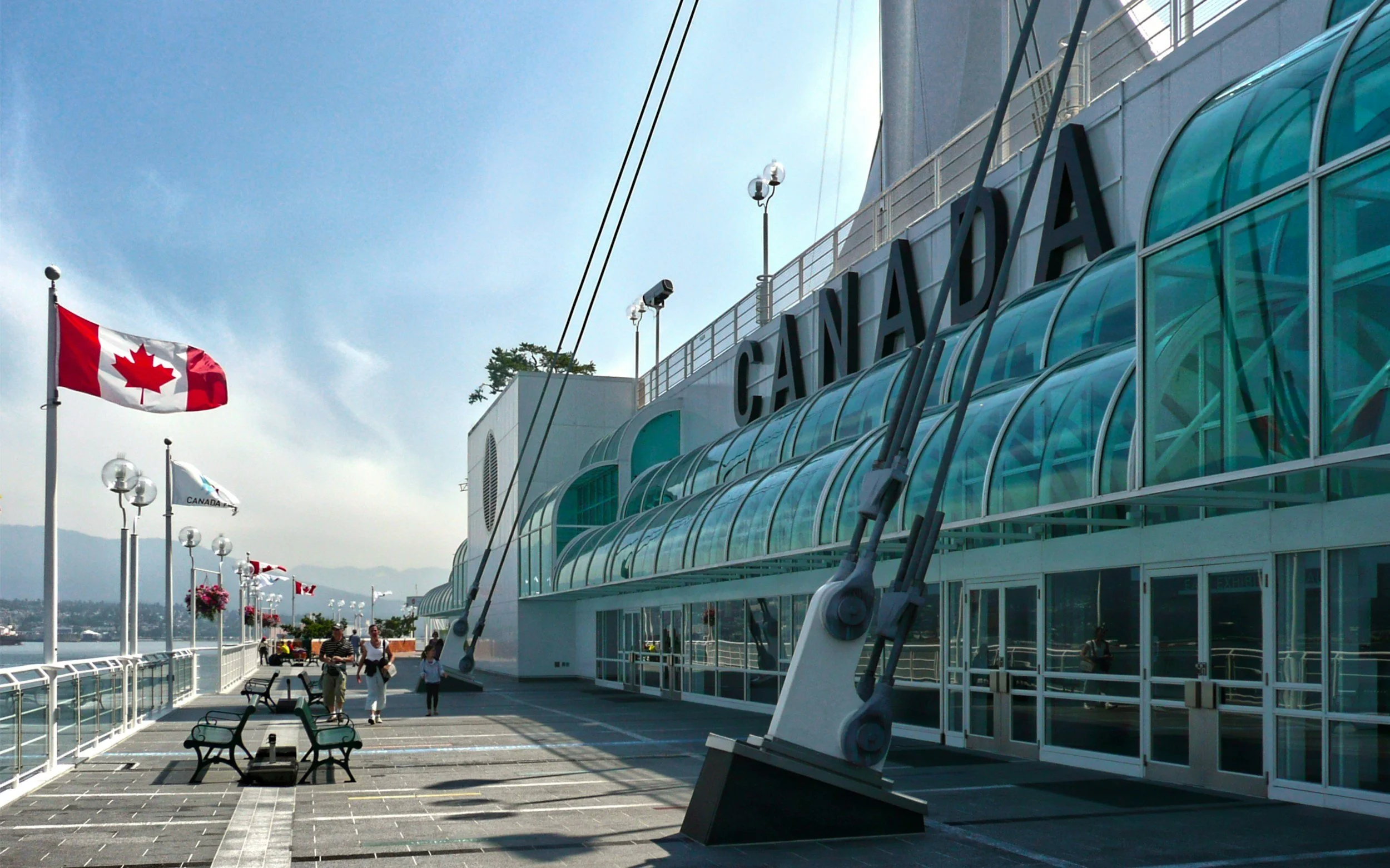 View of the Vancouver cruise ship terminal with flags, benches, and people walking along the waterfront pathway on a cloudy day.