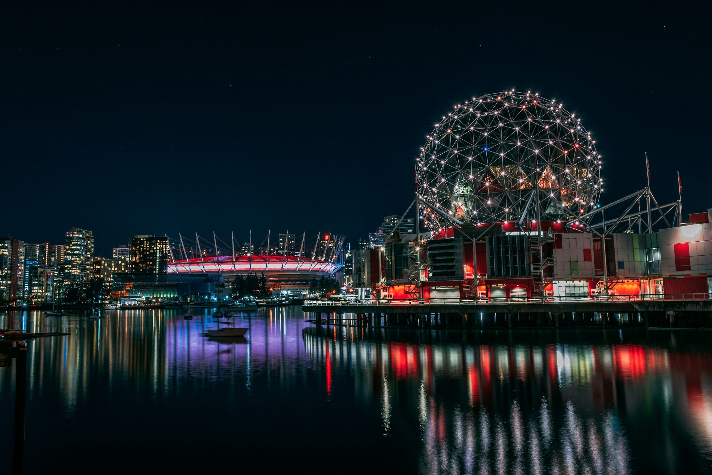 Night view of Vancouver skyline with Science World geodesic dome illuminated, reflecting on the water.