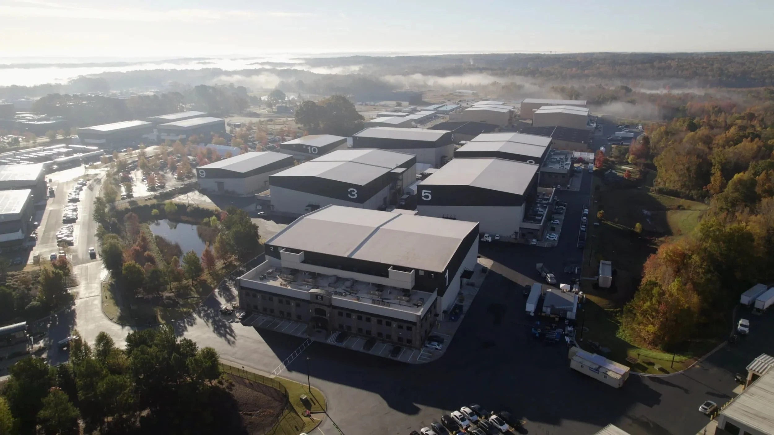 Aerial view of large industrial or warehouse buildings with numbered labels, surrounded by parking lots, trees, and a small pond, with a foggy horizon in the background.