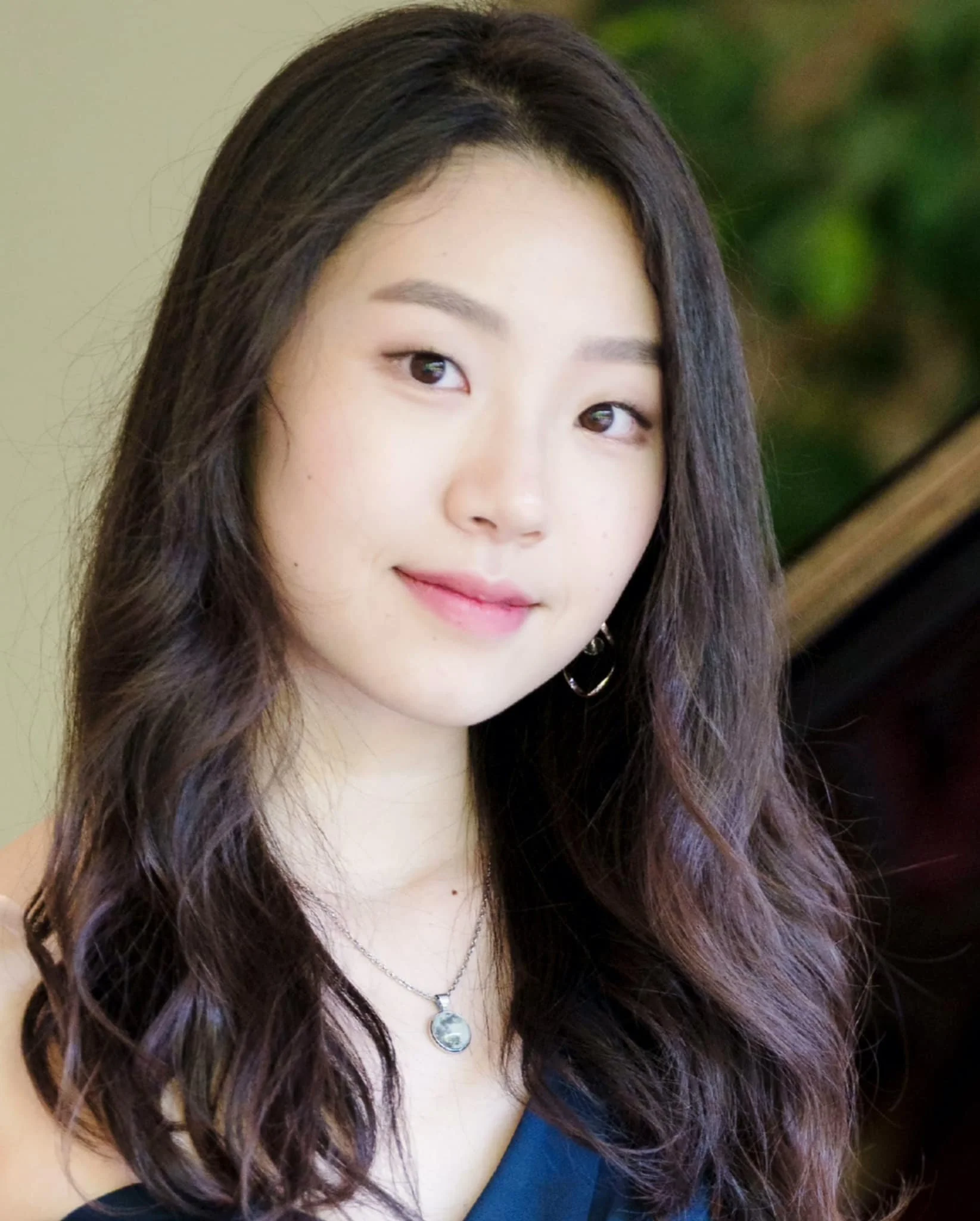 A young woman with long, wavy dark hair and light makeup, smiling gently, wearing a silver necklace with a circular pendant and a black top.