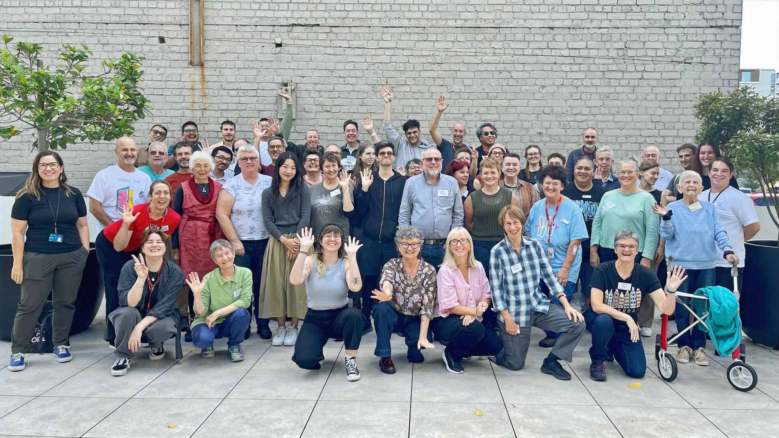 Group of diverse people smiling and waving outdoors in front of a brick wall, with green trees on either side.