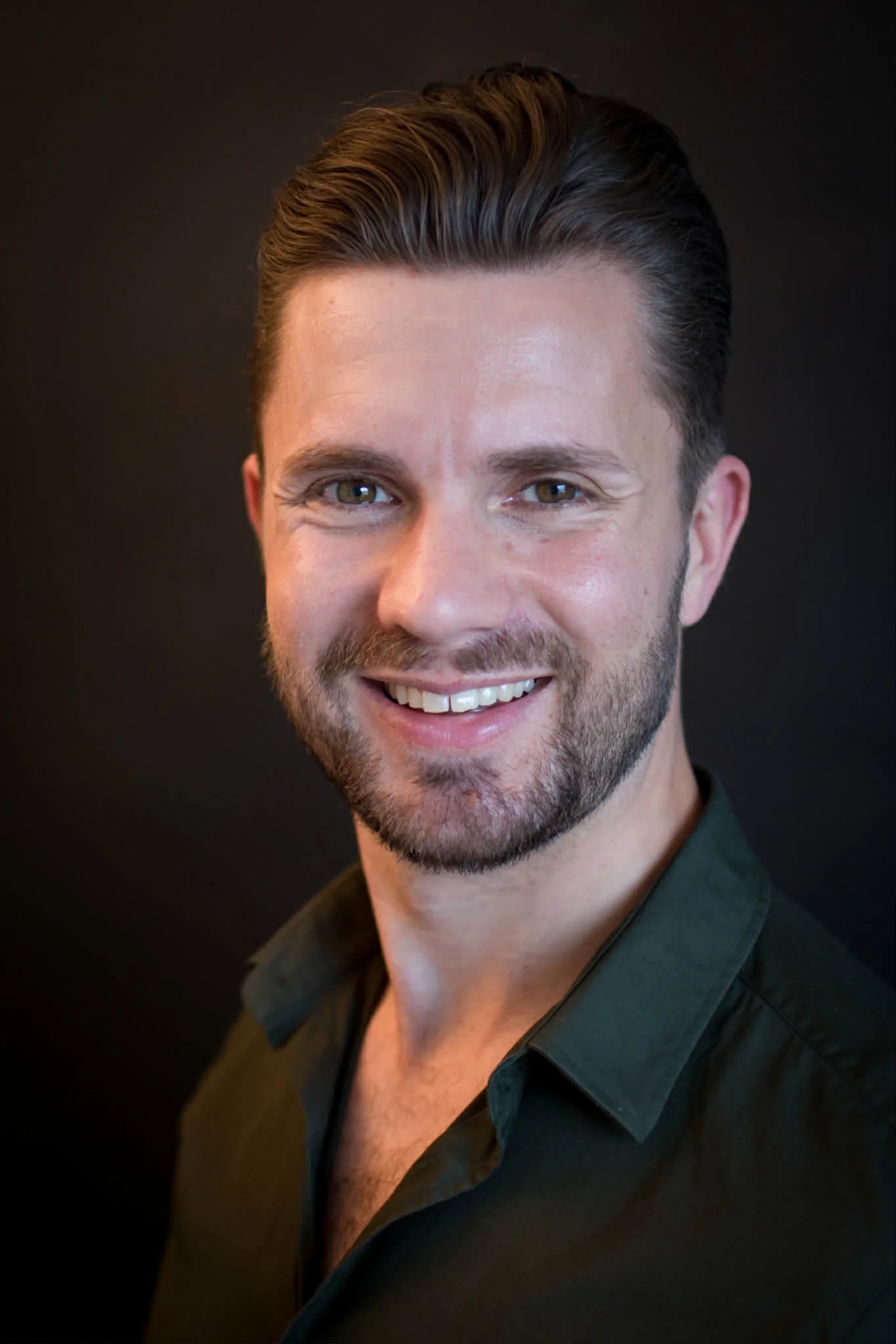 Close-up portrait of a smiling man with short dark hair, beard, wearing a dark green shirt, against a dark background.