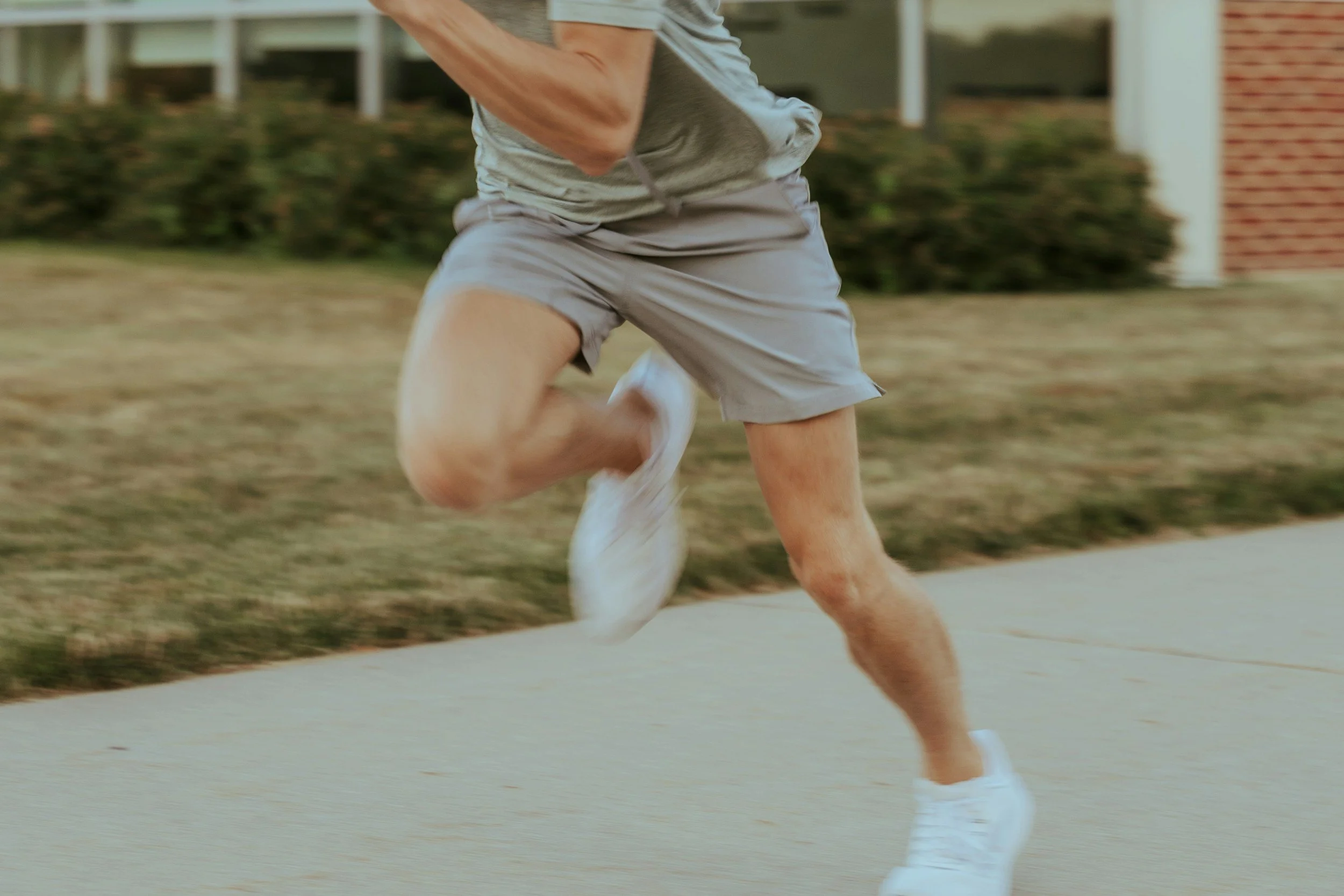 A person running on a sidewalk, wearing gray shorts, a gray t-shirt, and white running shoes, with a house and grass in the background.