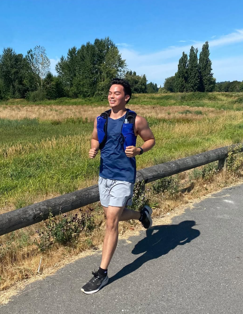 A young man jogging on a trail beside a grassy field with trees under a blue sky.