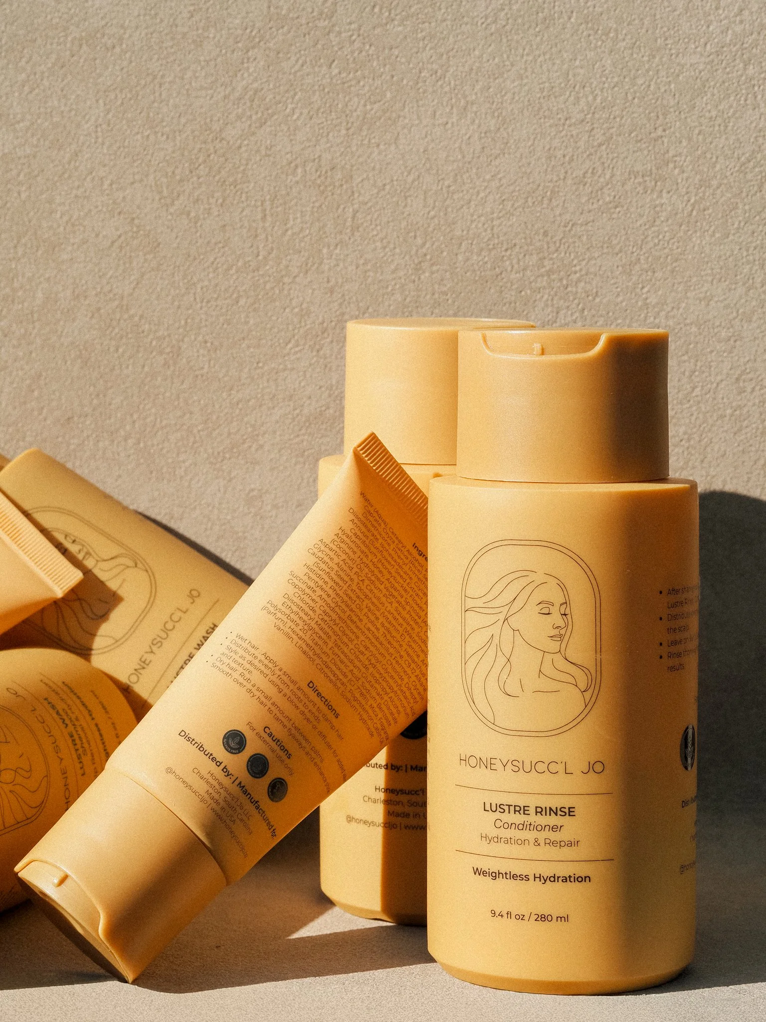 Multiple bottles of HONEYSUCCL JO hair care products with yellow packaging and minimalist line art logo, against a neutral background.