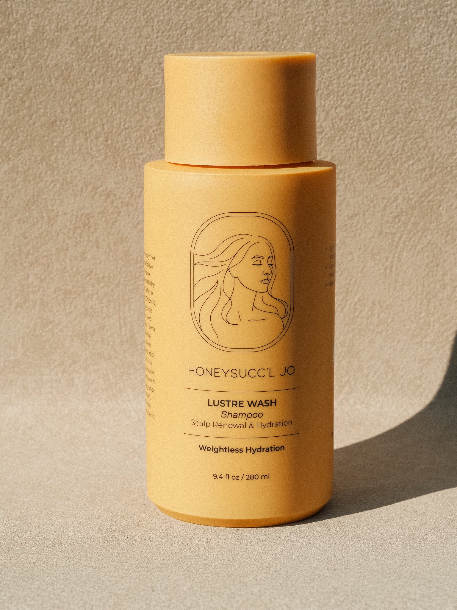 A yellow bottle of HoneySuck'l Jo Lustre Wash Shampoo for scalp renewal and hydration, with a line drawing of a woman's face and flowing hair on the label, placed on a neutral surface with a light-colored background.