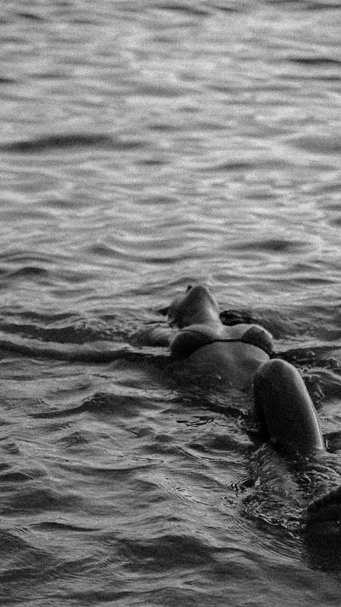 Black and white photo of a person floating on their back in water, with only their face and arms visible.