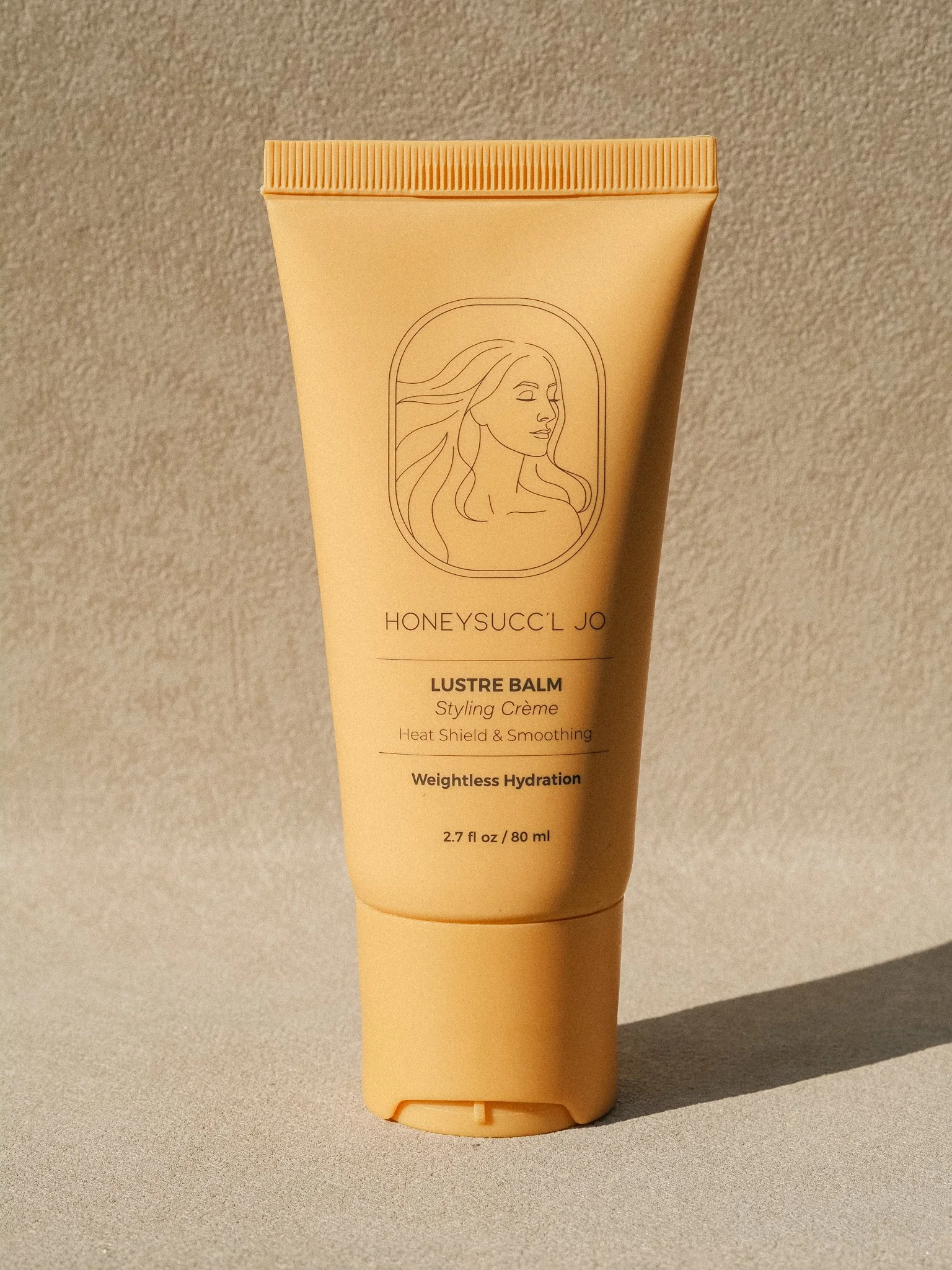 A peach-colored tube of HONEYSUCC'L JO Lustre Balm styling crème with a minimalist line drawing of a woman with flowing hair on the front, standing on a beige surface with textured background.
