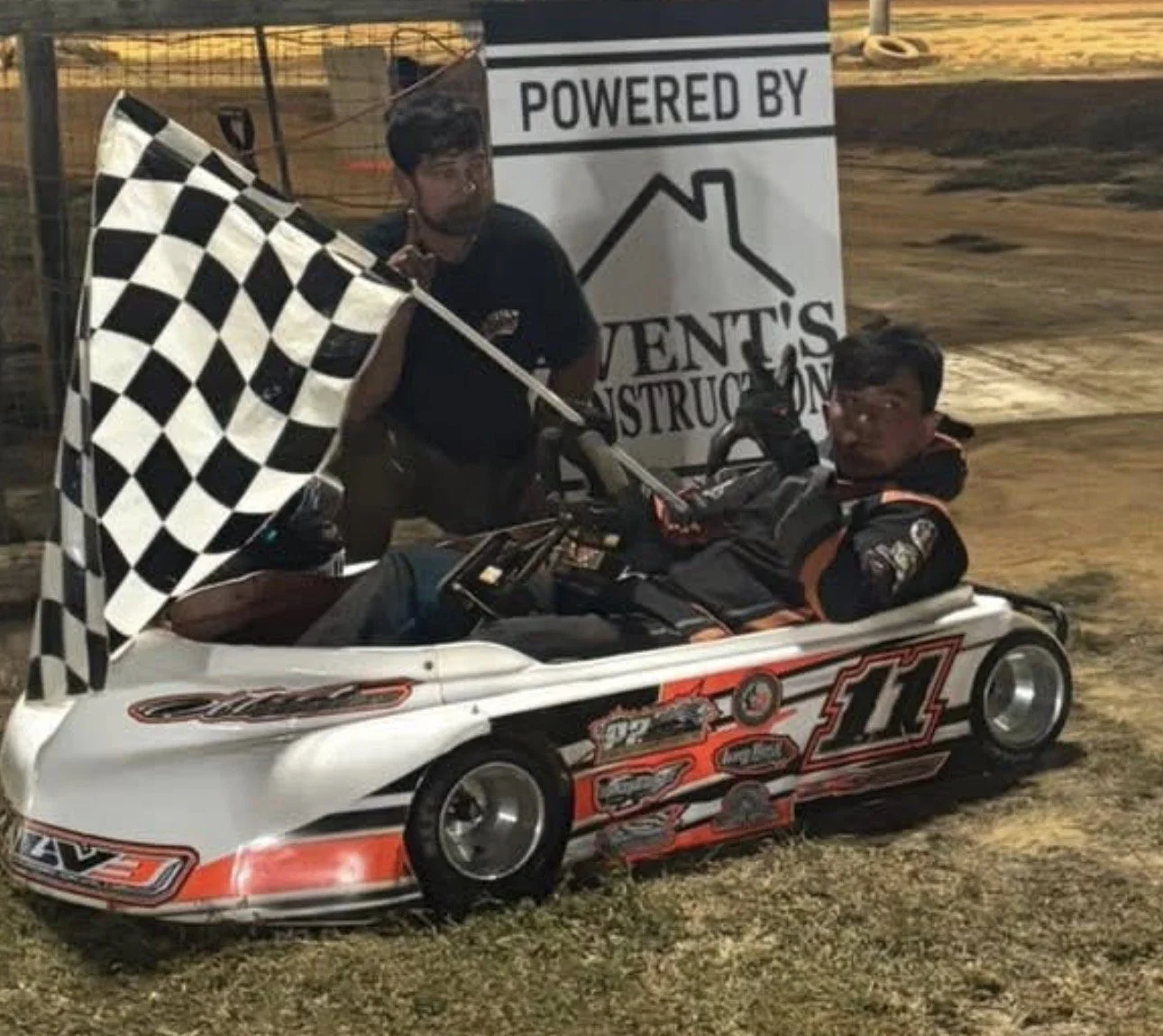 A young man in a racing suit sitting in a go-kart with the number 11 on it, holding a checkered flag. Next to him, a man standing with a black cap. They are at a dirt race track, with a sign in the background that reads 'Powered by Vent's Construction'.