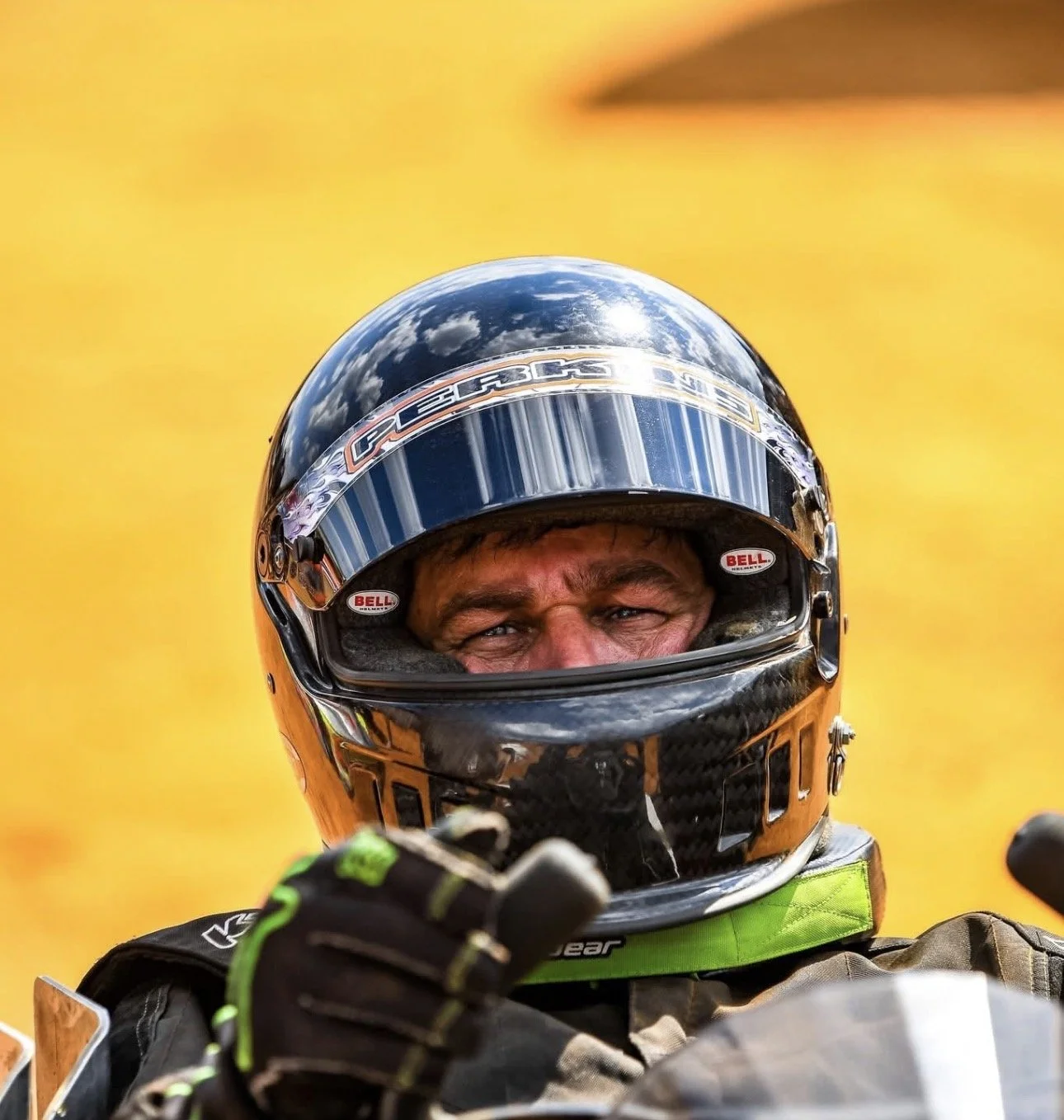 Close-up of a race car driver in a helmet, pointing at the camera with a yellow background.