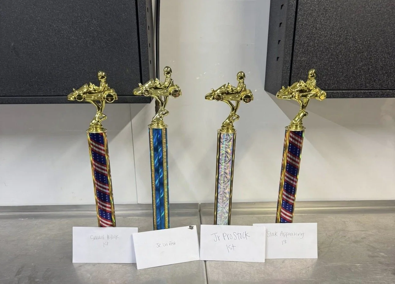 Four racing trophies with gold car and driver figure on top, colorful striped bases, and white handwritten labels in front of each.