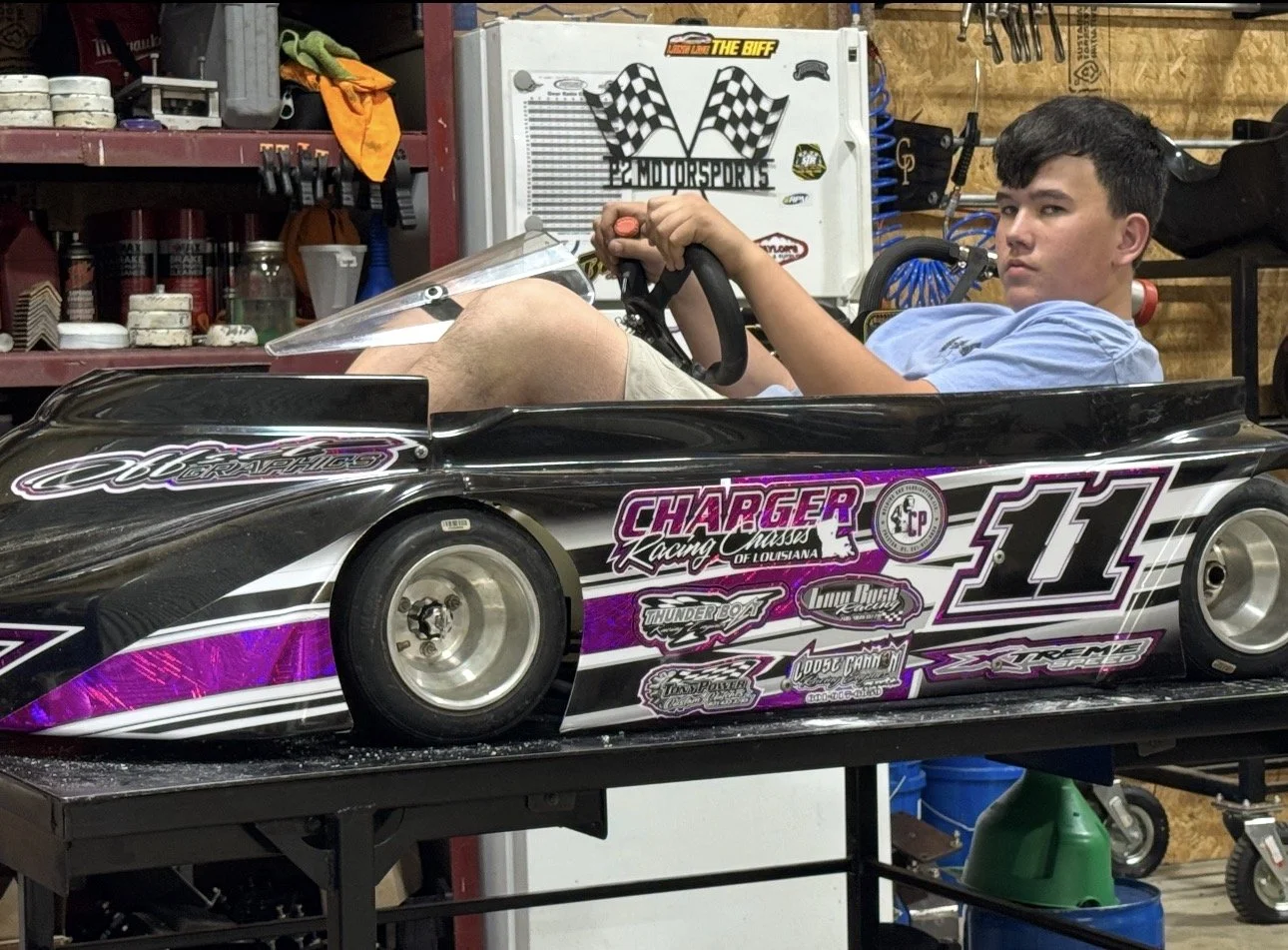 A young man sitting in a black and purple race car model in a garage with various tools and equipment around him.