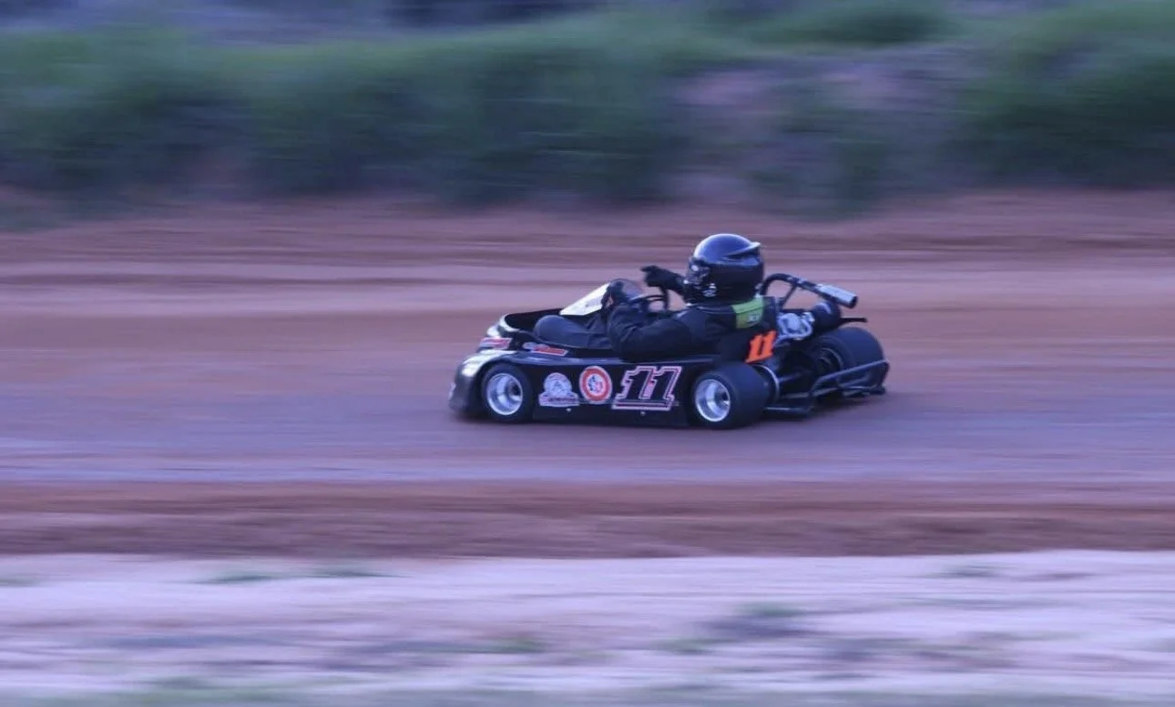 Person wearing a helmet and racing suit driving a go-kart on a dirt track.