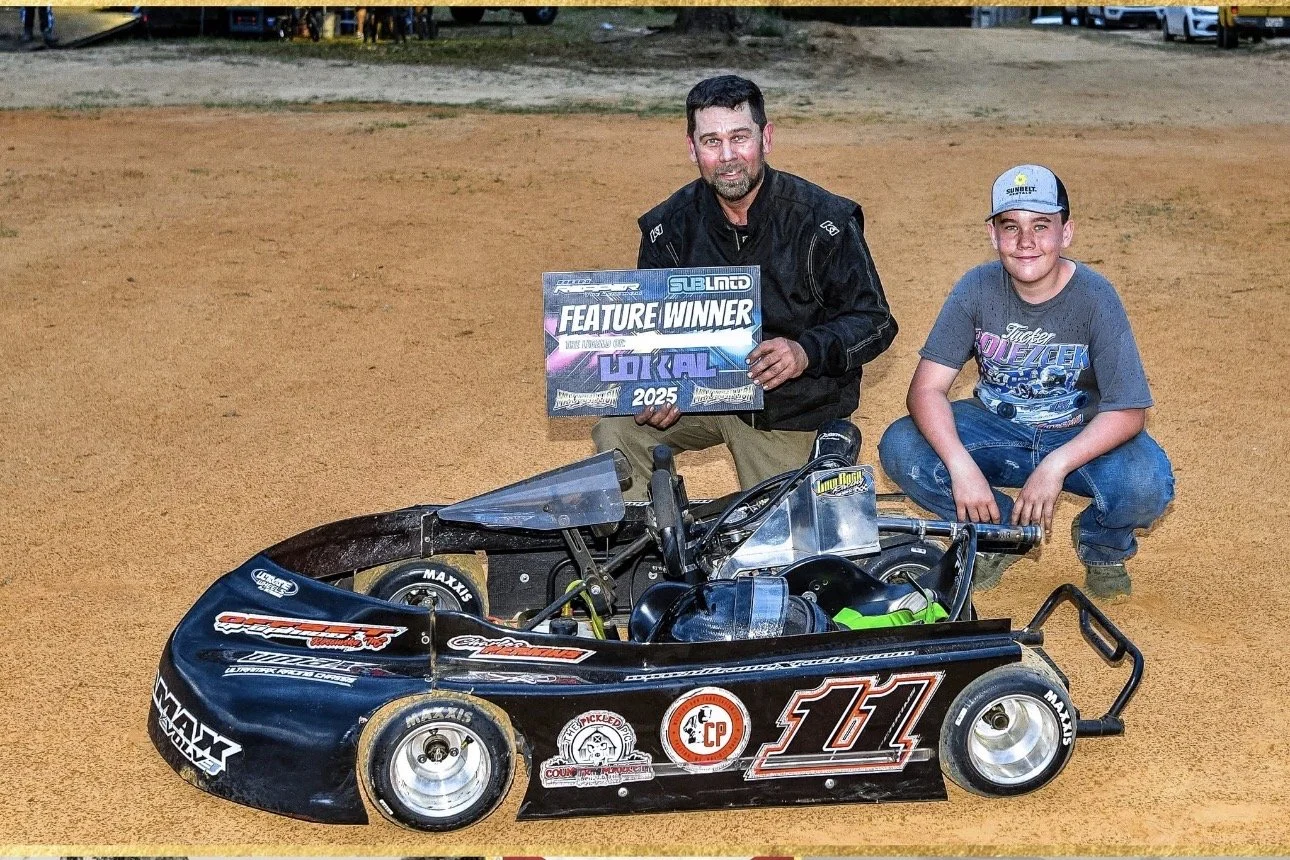 A man and a boy kneeling on a dirt race track next to a small race car, holding a sign that says 'Feature Winner' for the 2025 season, with the man wearing a black racing suit and the boy wearing a gray t-shirt and jeans.