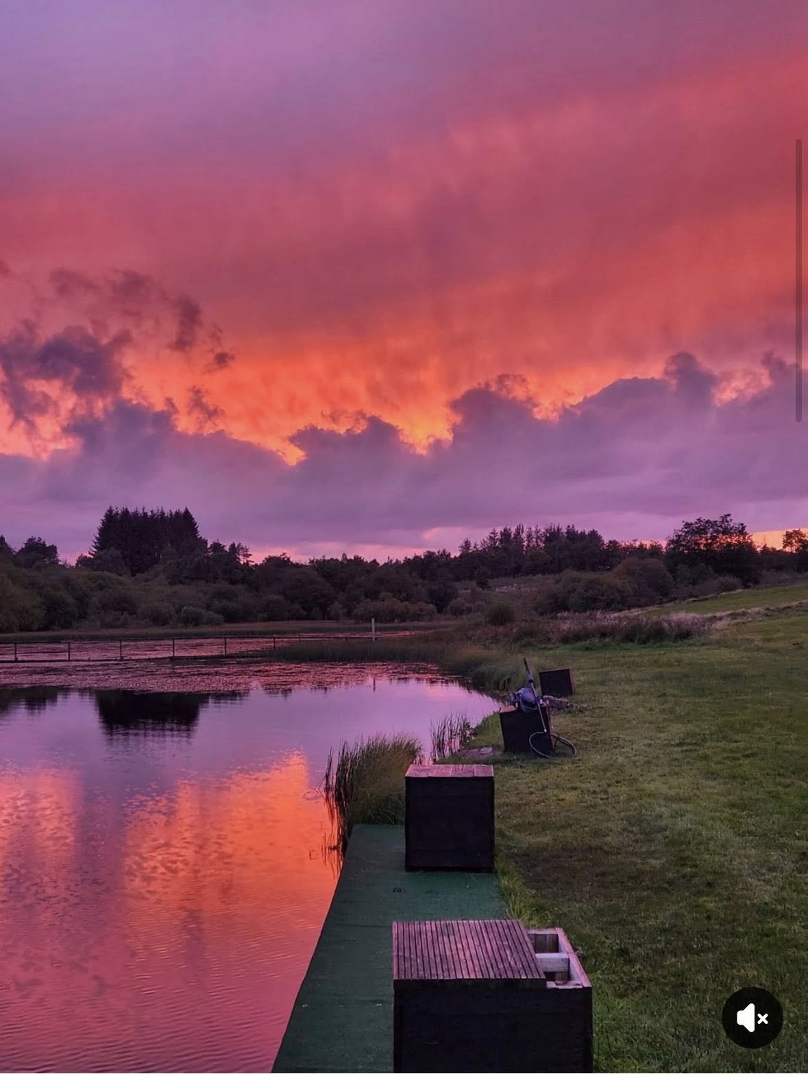 Sunset sky with vibrant pink, orange, and purple clouds above a calm river, with grass and bushes along the riverbank and some outdoor furniture on a green deck.