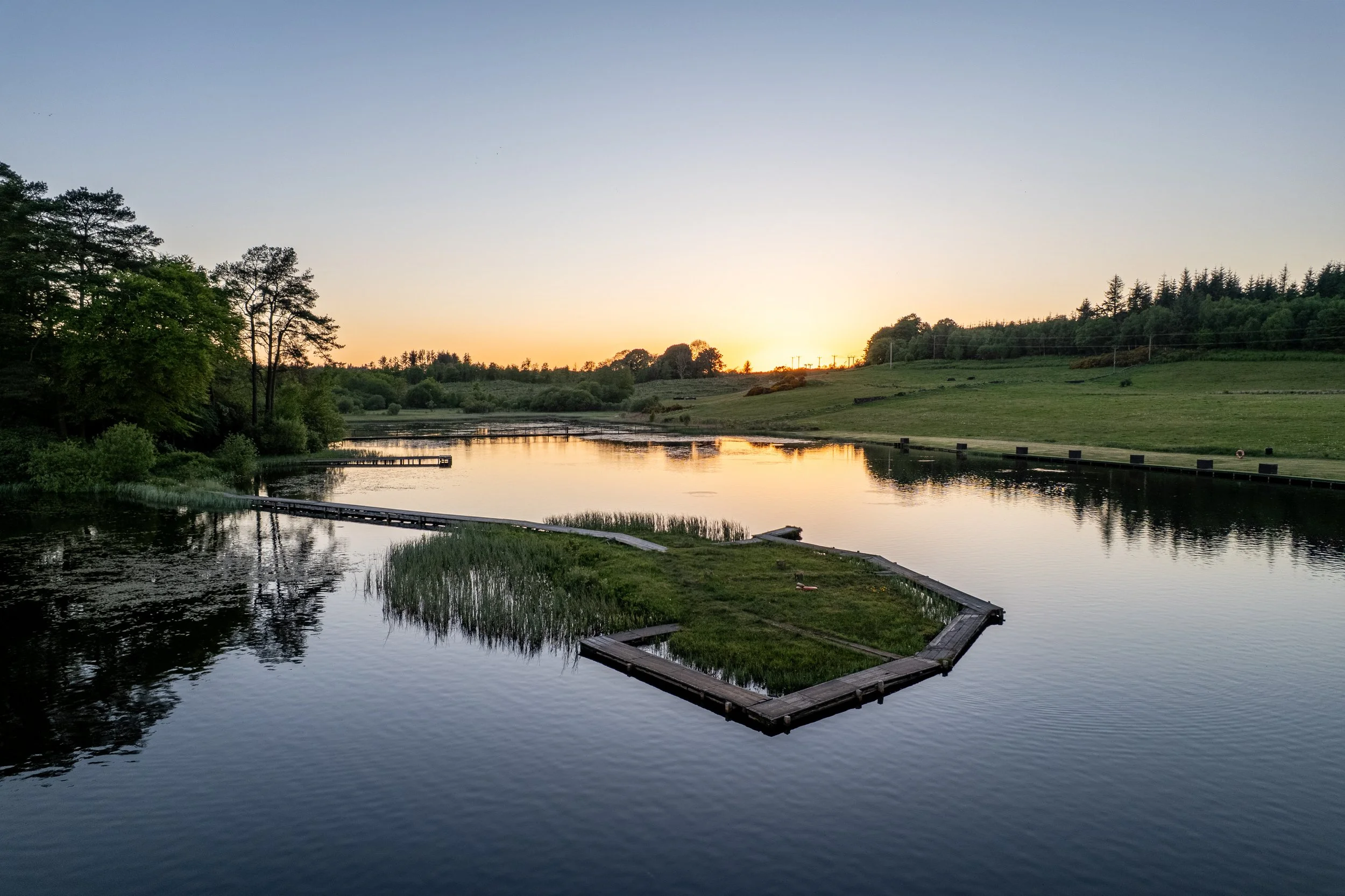 Calm river at sunset with greenery and trees on the banks, and a grassy island with a wooden fence.