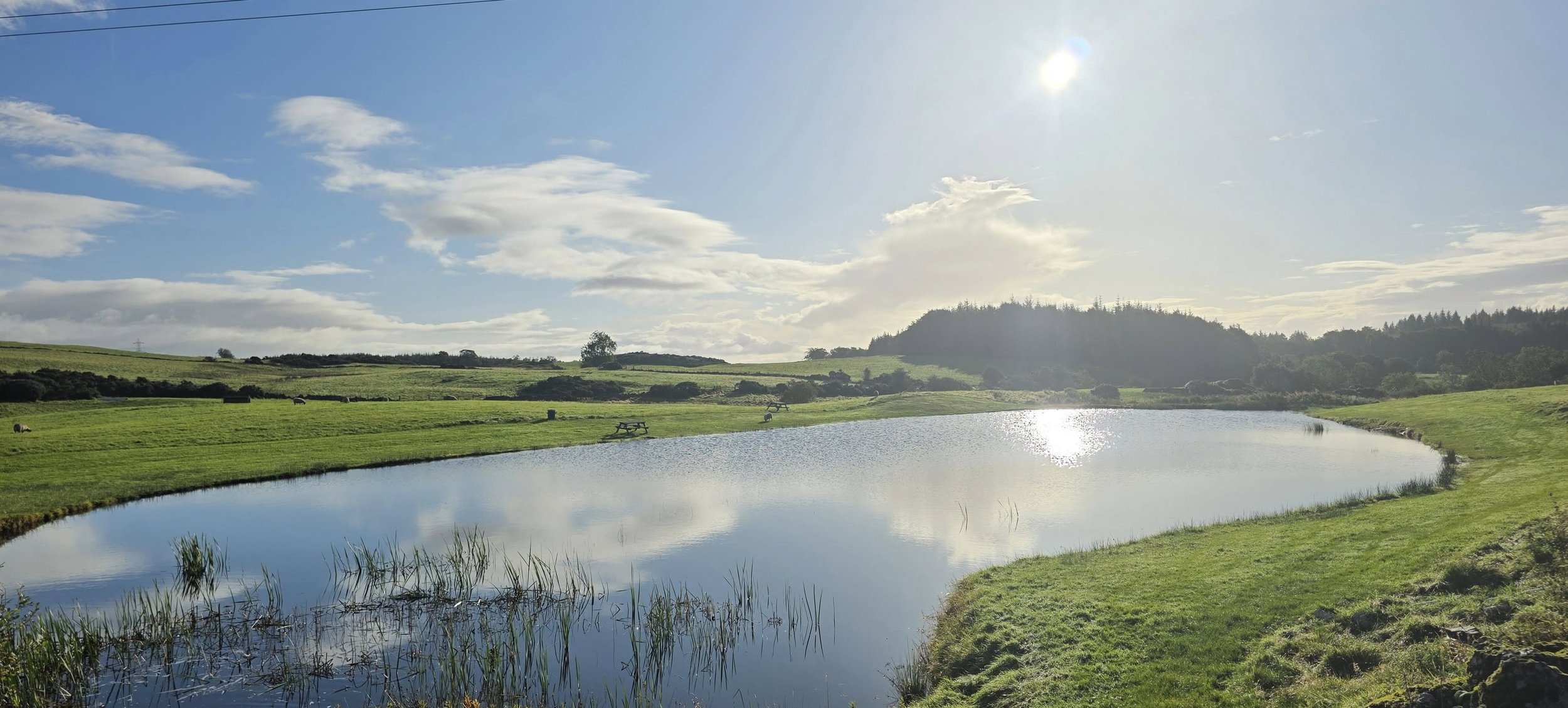 Scenic view of a small pond surrounded by green grassy fields, with hills and trees in the distance under a partly cloudy sky with the sun shining.