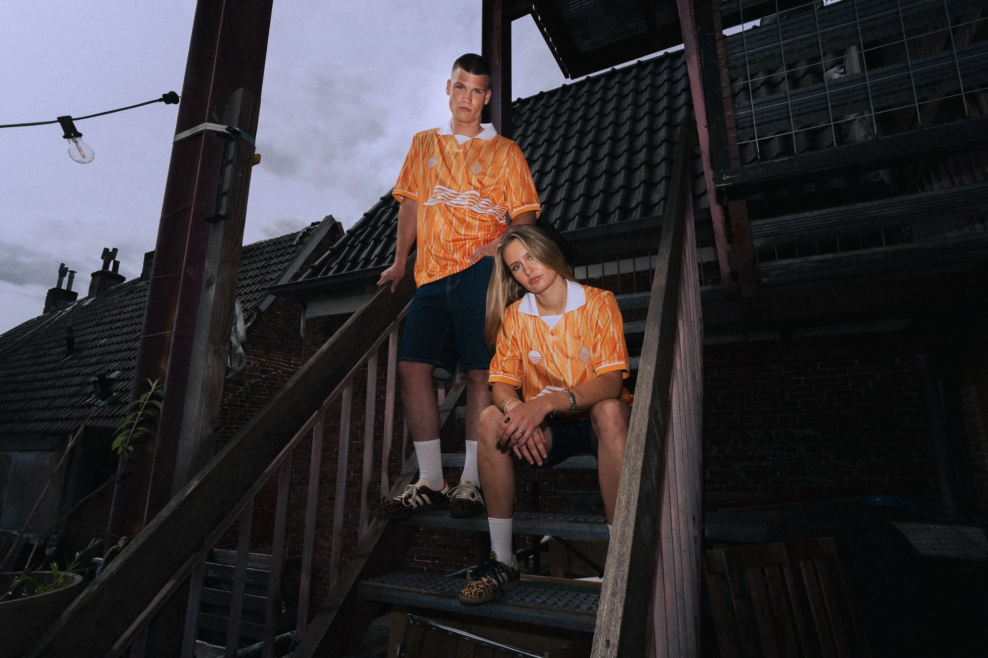Twee jonge mensen poseren op een buitentrap met een donkere, partly cloudy avondlucht op de achtergrond. Ze dragen matching oranje shirts met patroon en witte coltruien eronder.