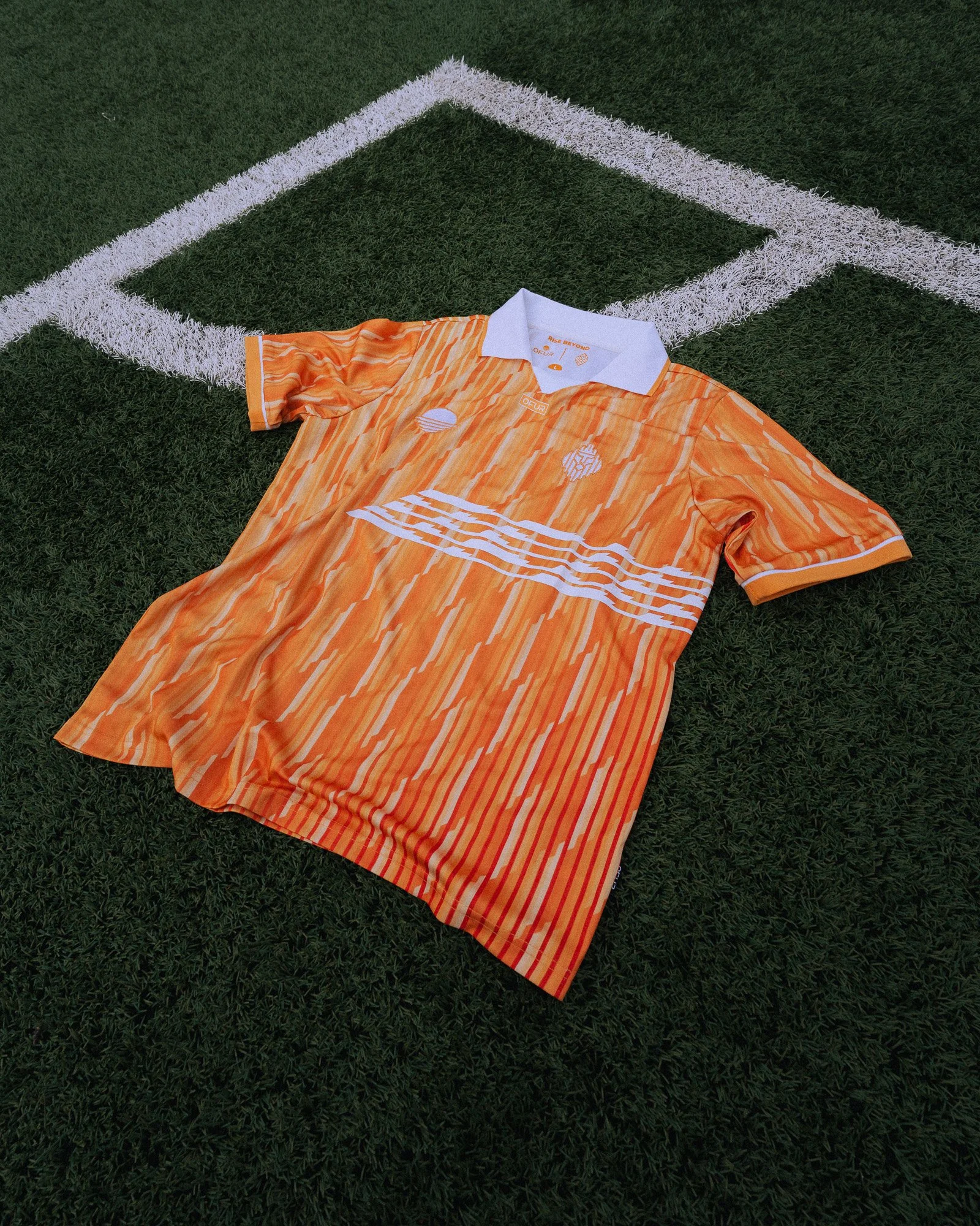 Oranje voetbalshirt met witte details ligt op kunstgrasveld met witte hoeklijn.