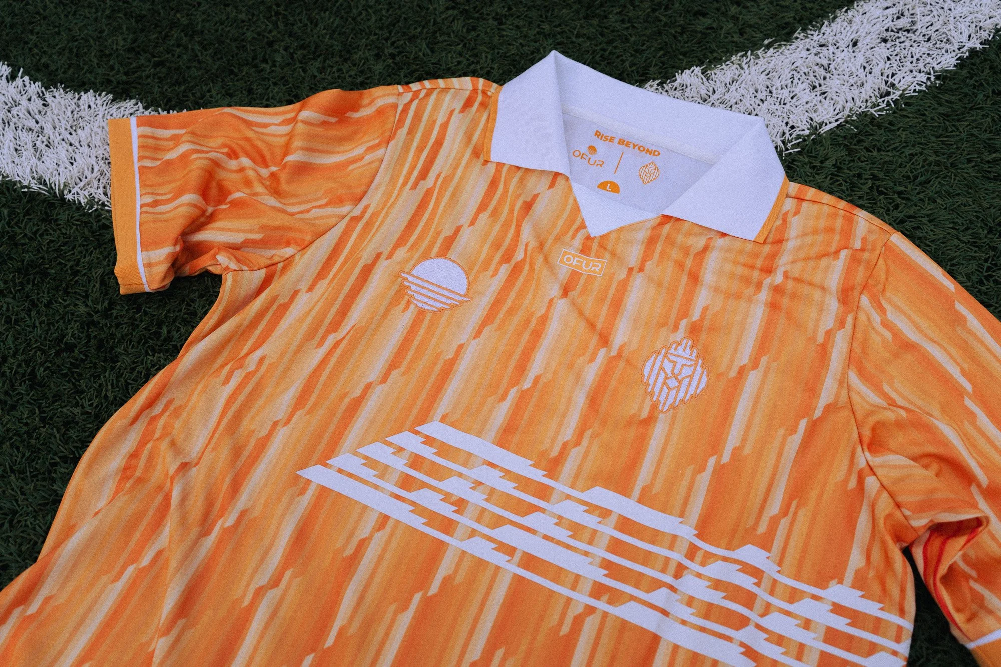 Een oranje sportshirt met witte en oranje strepen, ligt op gras. Het shirt heeft een witte kraag en heeft verschillende witte en oranje logo's en teksten, waaronder 'RISE BEYOND' en 'OFUR'.