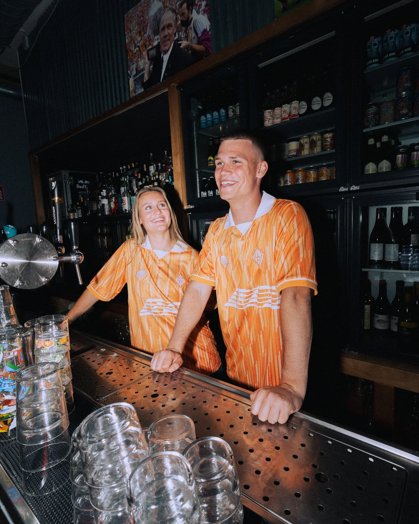 Twee mensen in oranje gestreepte shirts staan achter de bar in een café, lachend en pratend. Op de achtergrond bevinden zich verschillende alcoholflessen en gekoelde bier- en frisdrankblikken.