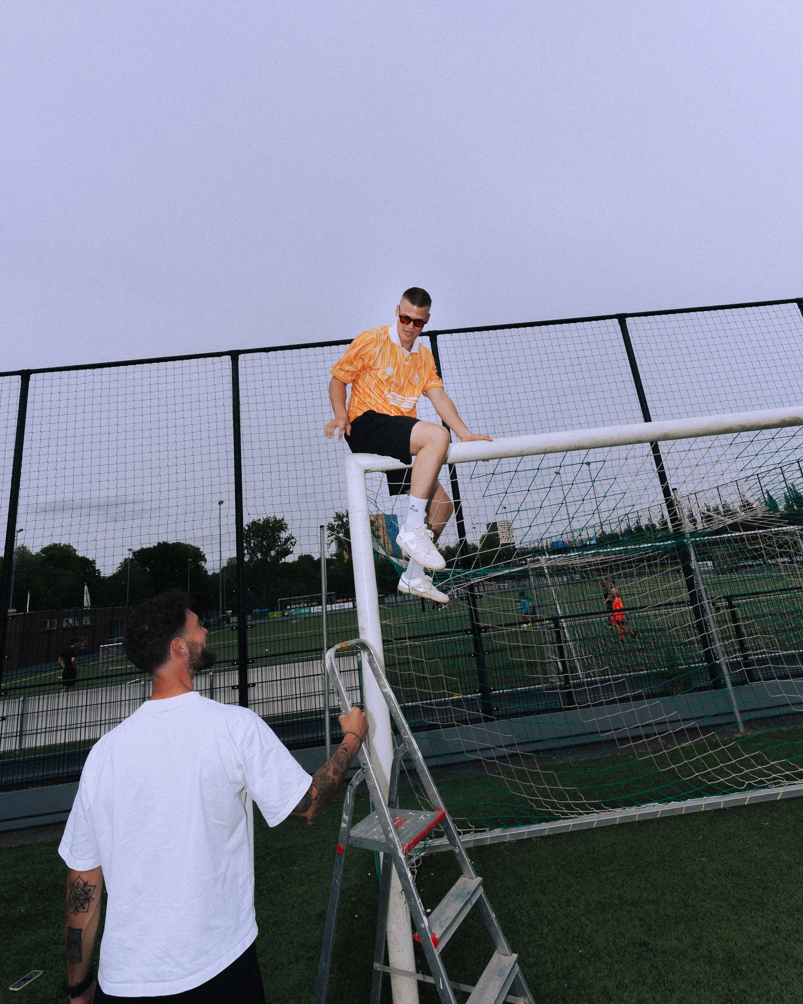 Een man met tattoos in een witte T-shirt kijkt terwijl een andere man hem op een ladder helpt om een FIFA World Cup billboard te bevestigen. Ze zijn op een grasveld met een voetbalveld op de achtergrond.