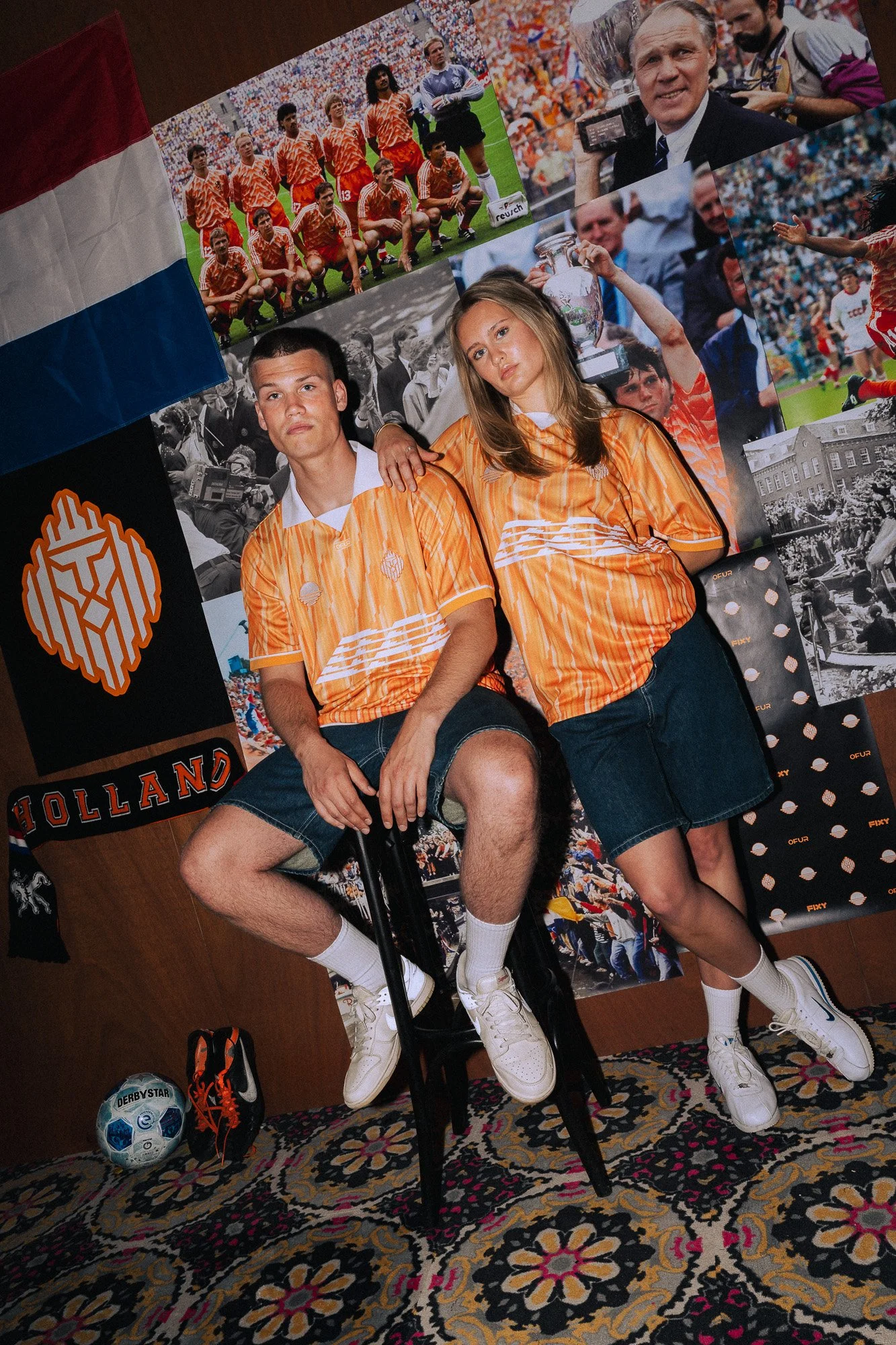 Twee jonge mensen in oranje sportkleding zitten op een stoel, omringd door foto’s en memorabilia van het Nederlands voetbalteam. Op de vloer liggen een voetbal en sportschoenen, op de achtergrond posters van een voetbalwedstrijd en teamfoto’s.
