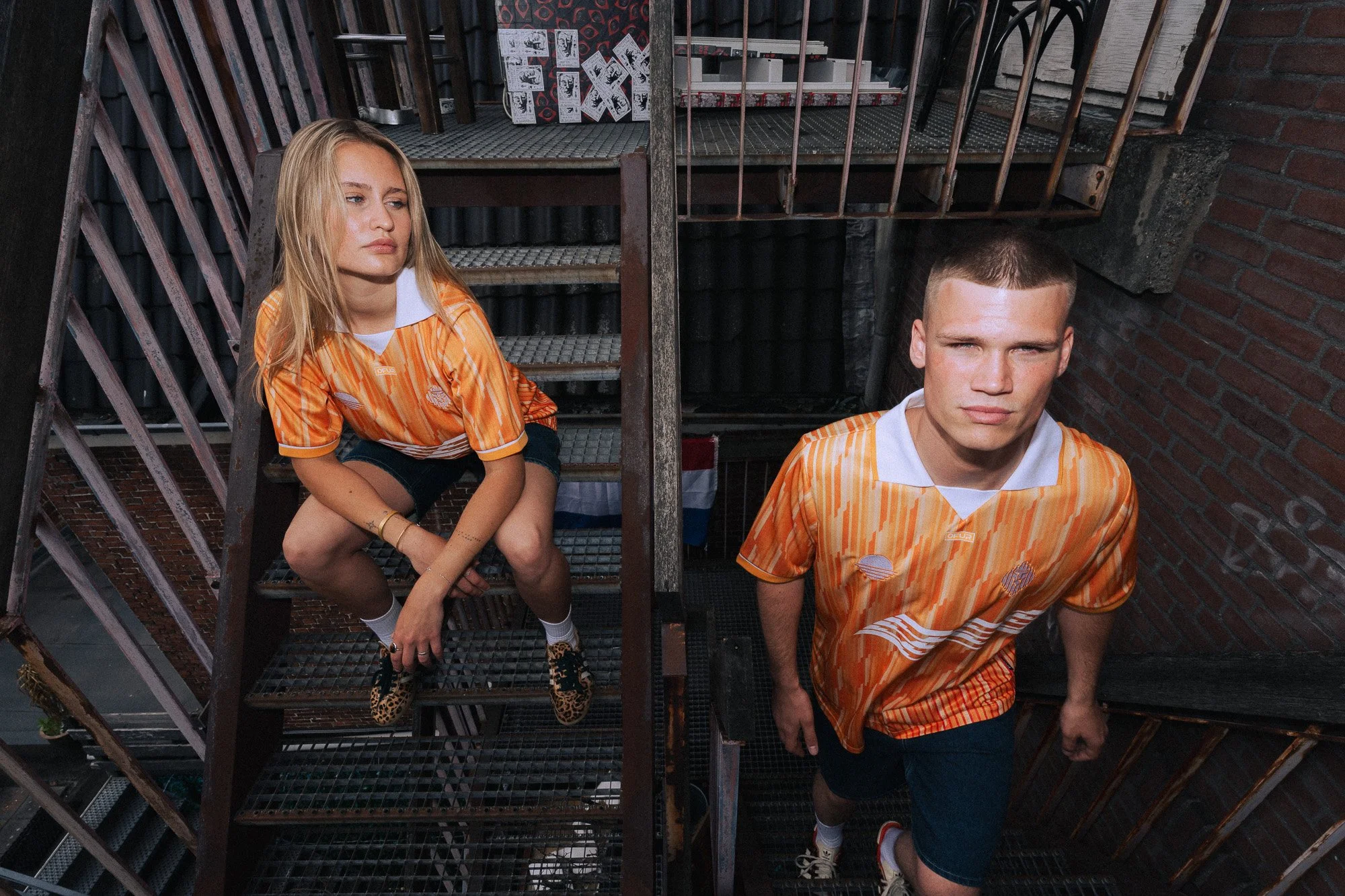Twee jonge mensen in oranje sportkleding poseren op een metalen brandtrap tegen een bakstenen muur, met posters en dozen op de achtergrond.