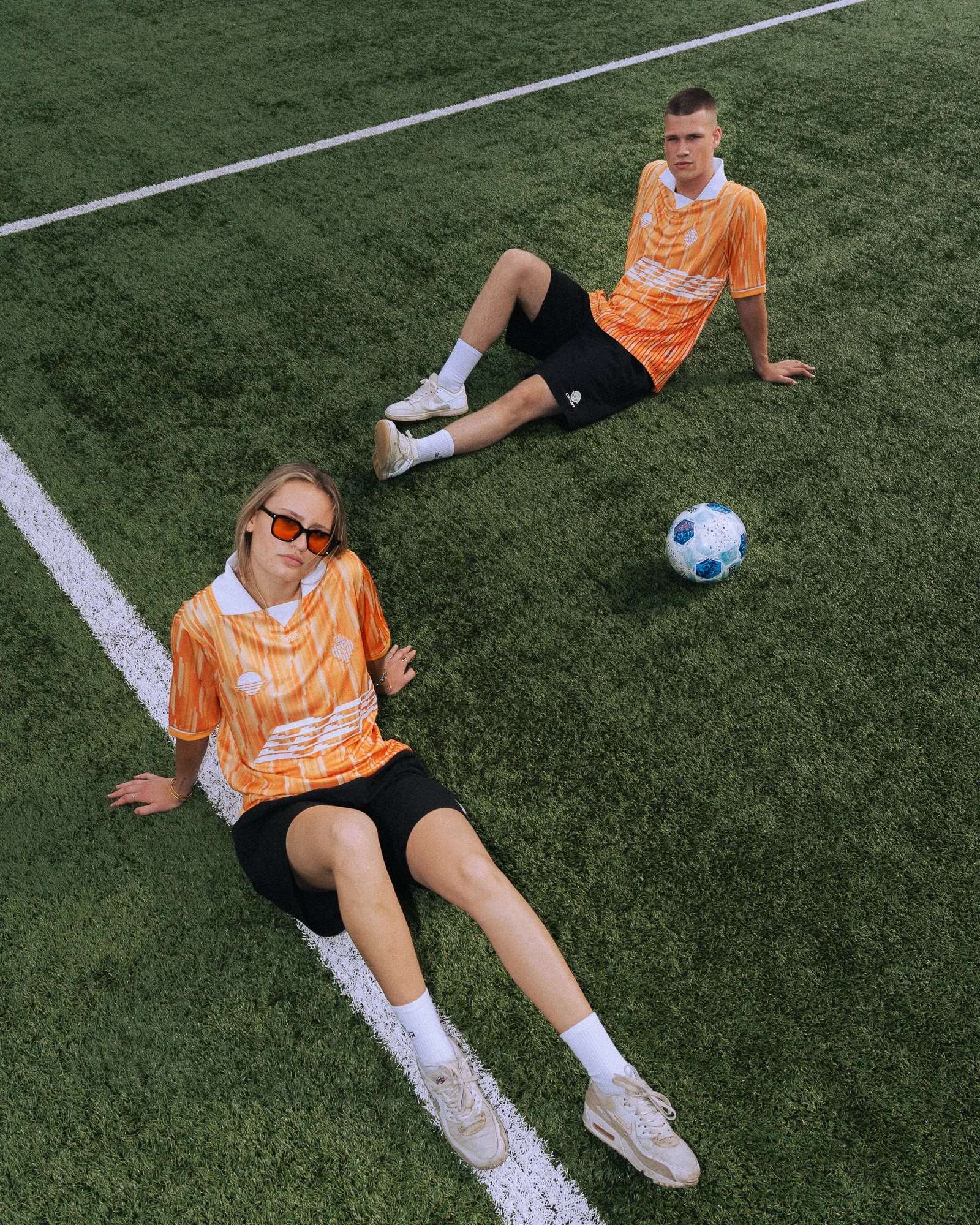 Twee jonge mensen in oranje sportkleding zitten op een voetbalveld, met een voetbal tussen hen.