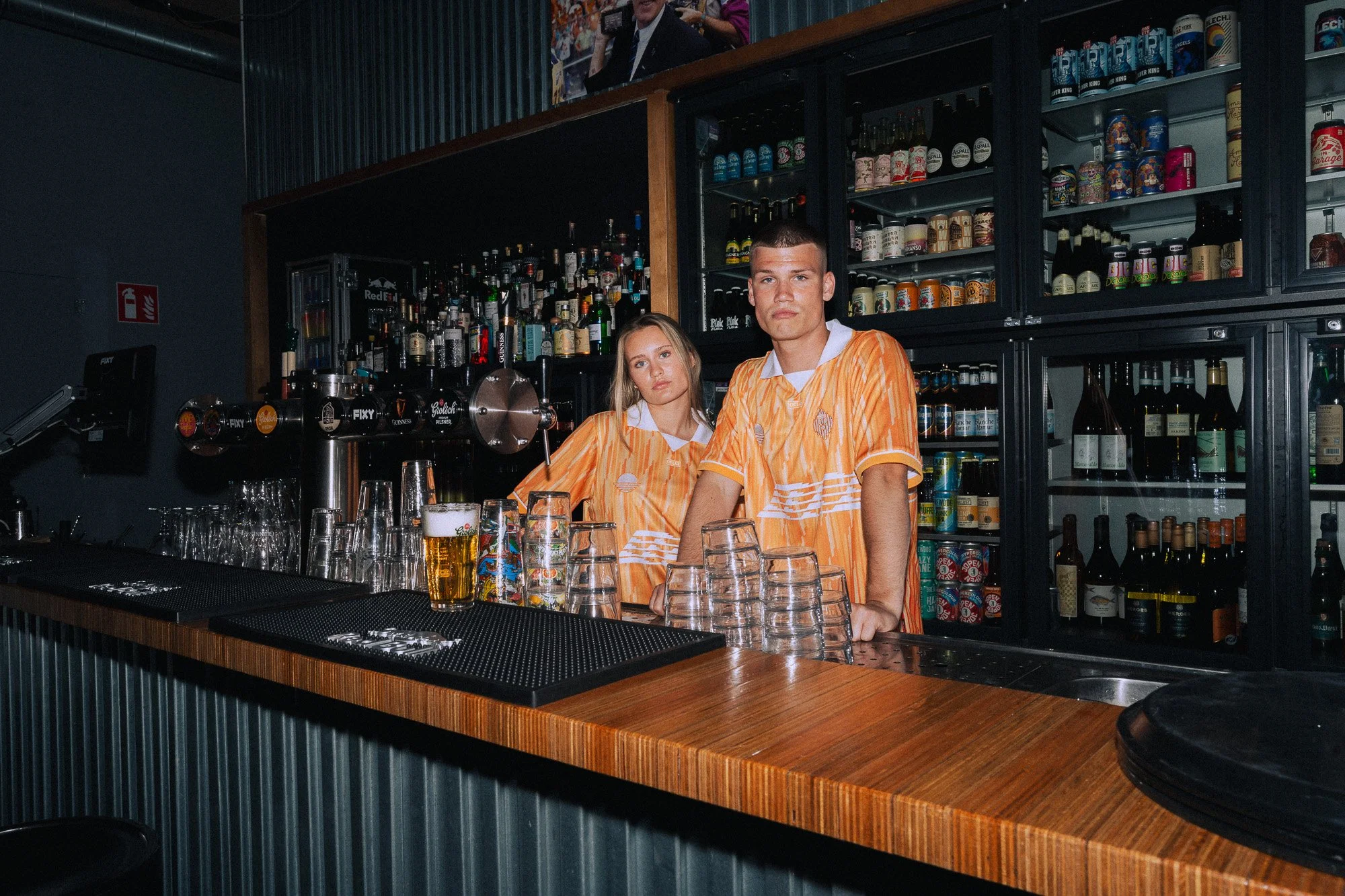 Twee jonge mensen in oranje gestreepte shirts achter de bar in een bar of café, omringd door glazen en flessen drank. Achter hen een muur vol met verschillende flessen alcohol en een koelkast met andere drankjes.