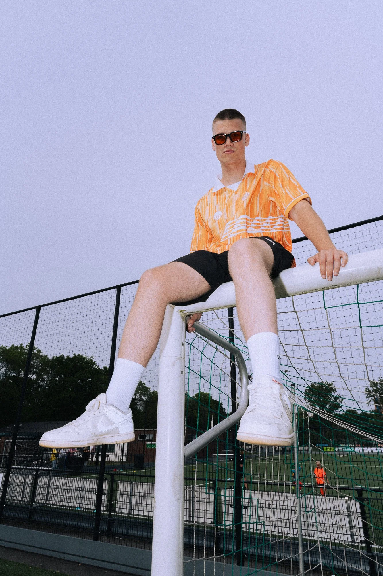 Jongeman met zonnebril zittend op een voetbaldoel, gekleed in een oranje shirt, zwarte shorts, witte sokken en sneakers op een sportvelden.
