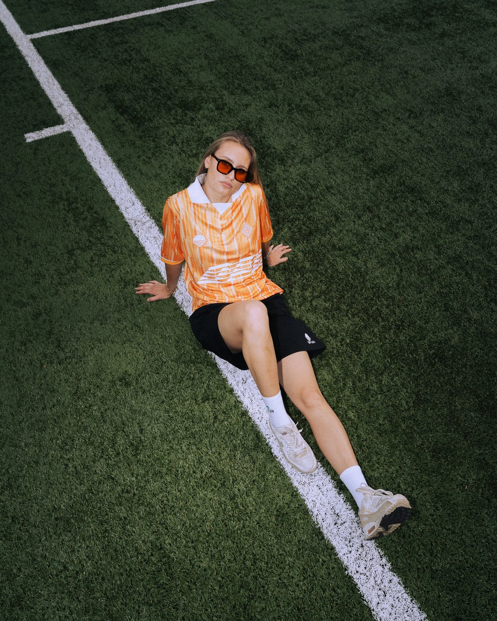 Een vrouw met witte sportkleding en oranje overhemd zit op een groen sportveld, leunend tegen een witte lijn.