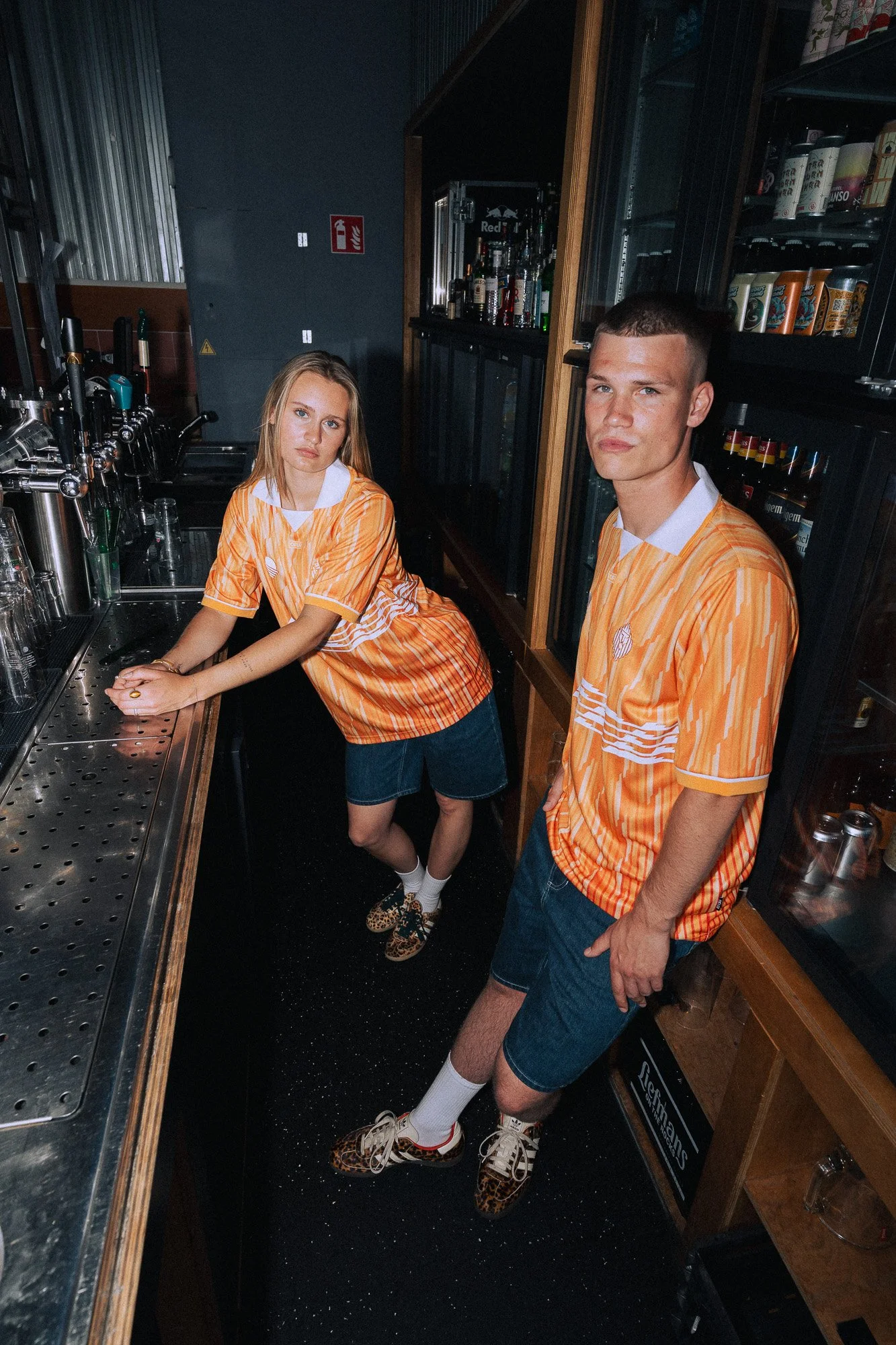 Twee jonge mensen in een bar, gekleed in oranje en wit geruite shirts, met een barachtergrond met drankflessen en glazen.